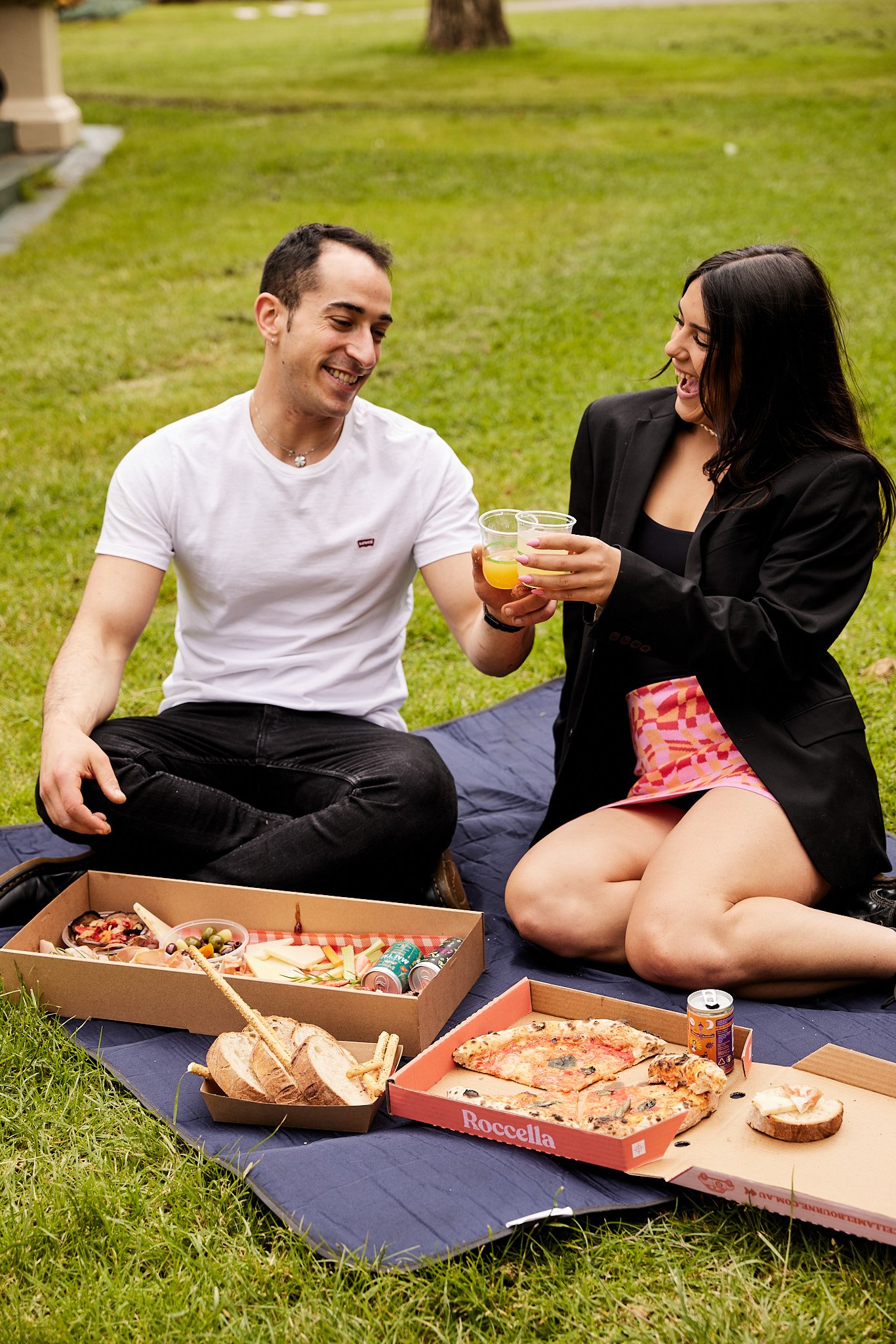 In Fitzroy Gardens, two people are enjoying a delightful afternoon with Curatif cocktails and Roccella picnic boxes. The cocktails are expertly mixed, with bold and complex flavors that perfectly complement the delicious food. As they sit on a blanke