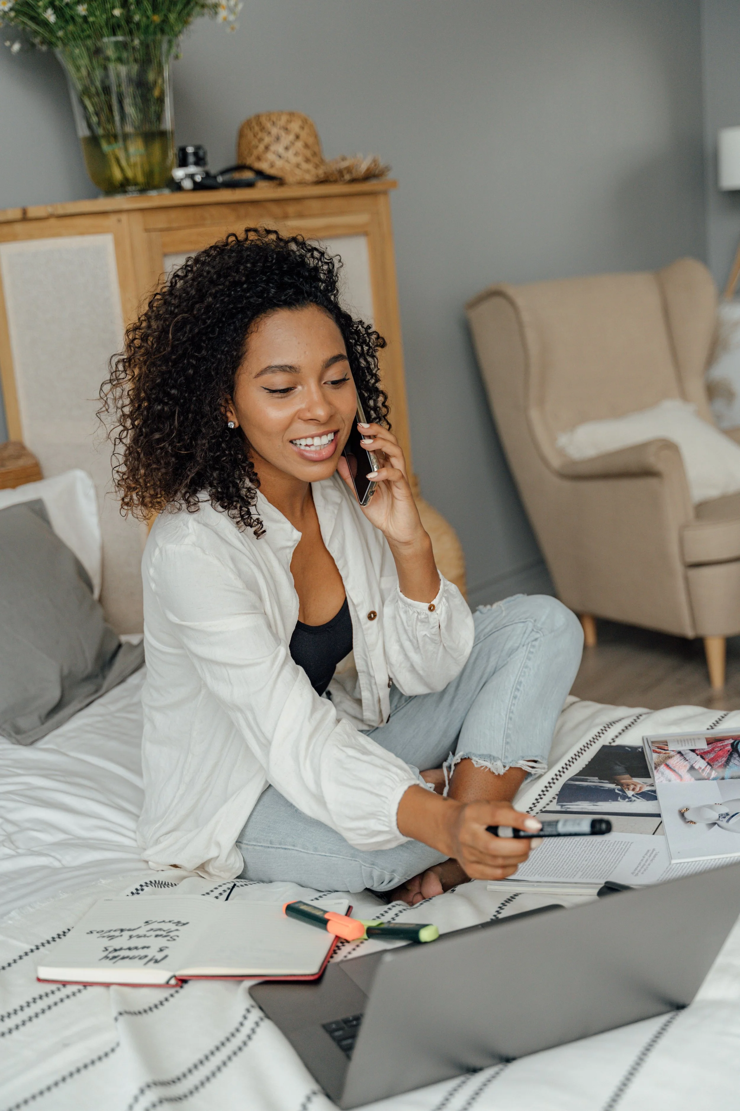 Woman sitting on bed, talking on phone, using a laptop, with open notebook and highlighters nearby.