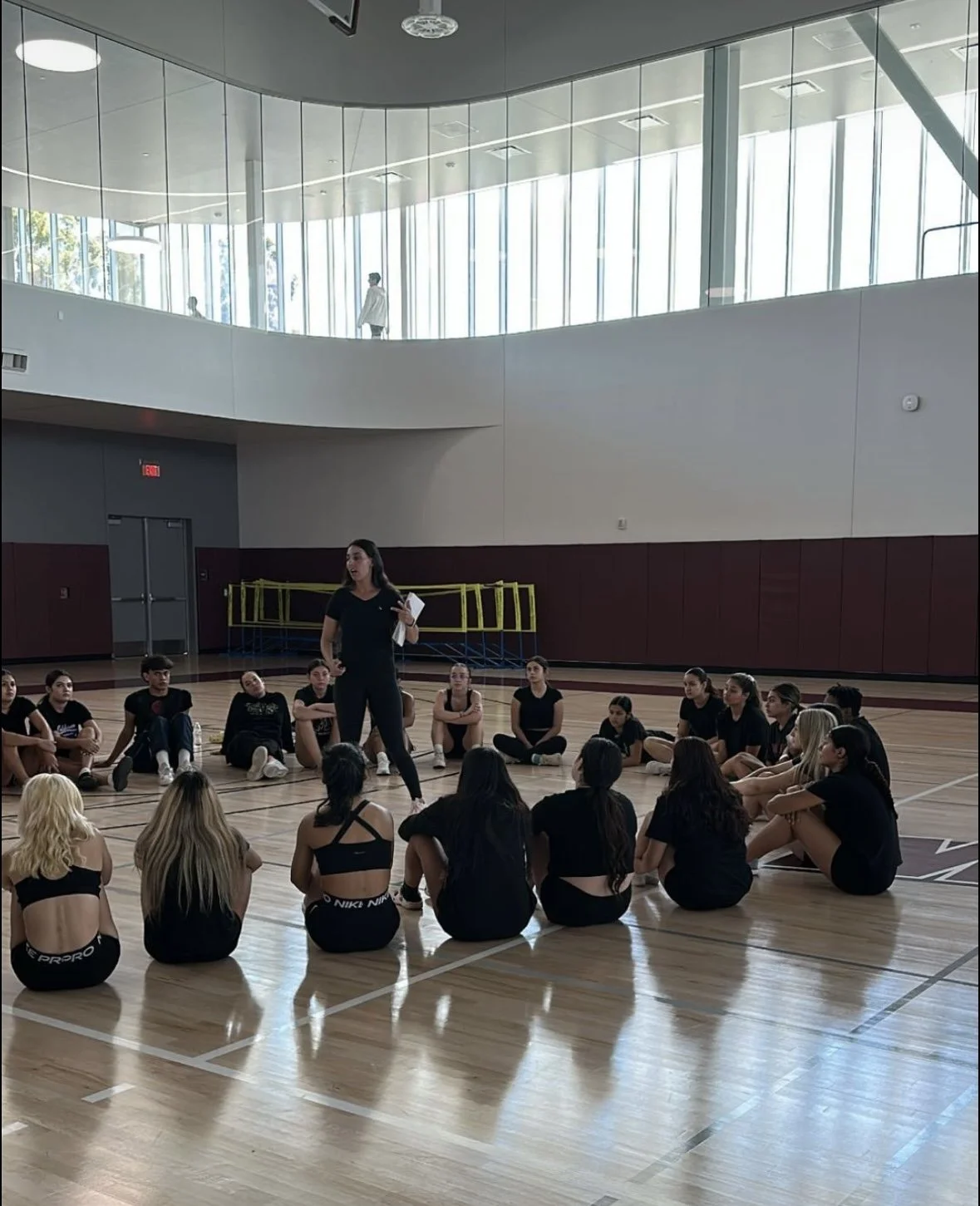 A group of young women sitting on the gymnasium floor in a circle, with a woman standing in the center speaking to them. The women are dressed in athletic wear. There are large windows in the background and a balcony with a person walking above.
