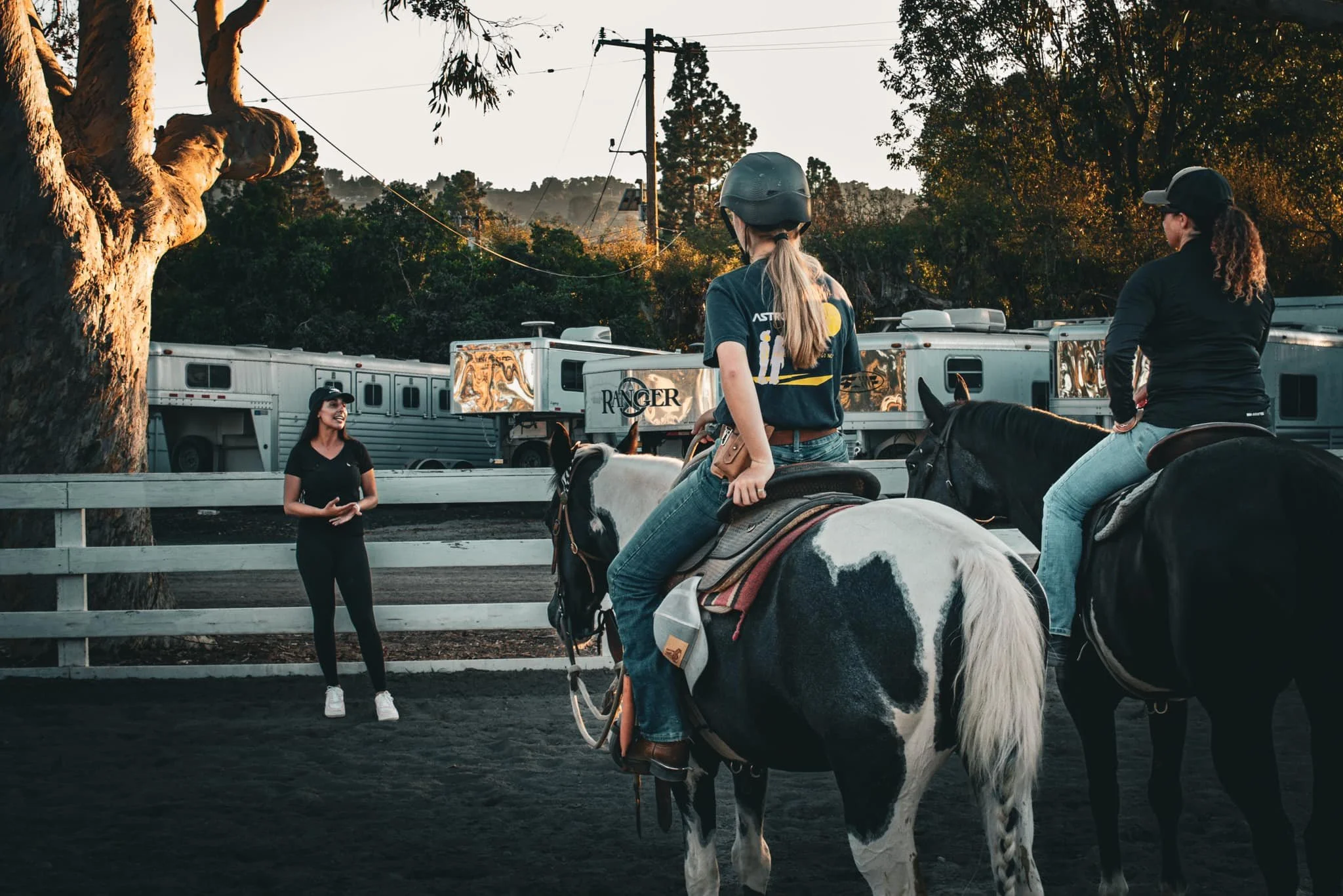 Two women on horseback listening to a woman in black speaking in an outdoor arena during the late afternoon, with trees and horse trailers in the background.