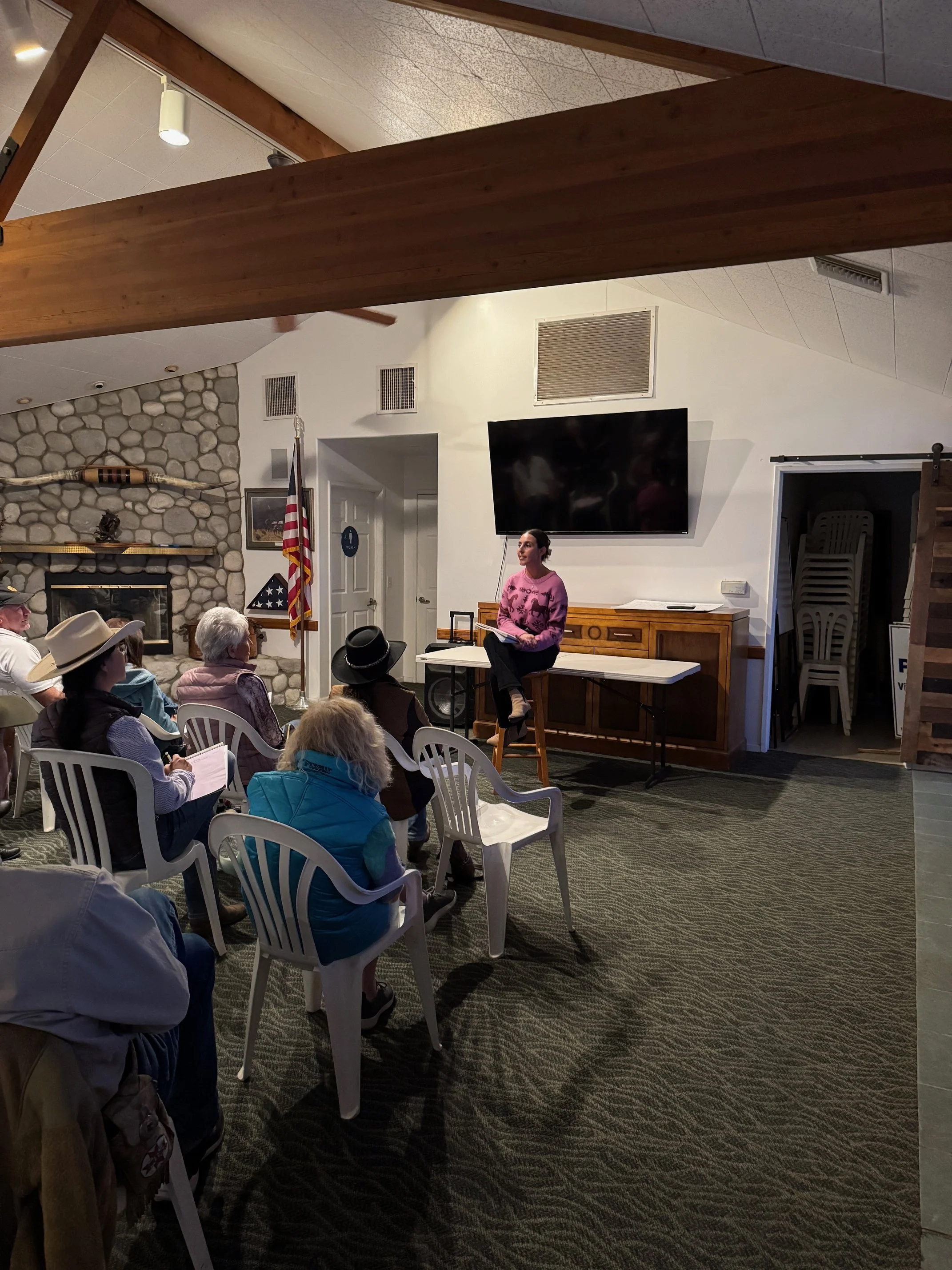 A woman in a pink sweater speaking to a group seated on white plastic chairs in a cozy indoor setting with a stone fireplace, American flag, and large television screen.
