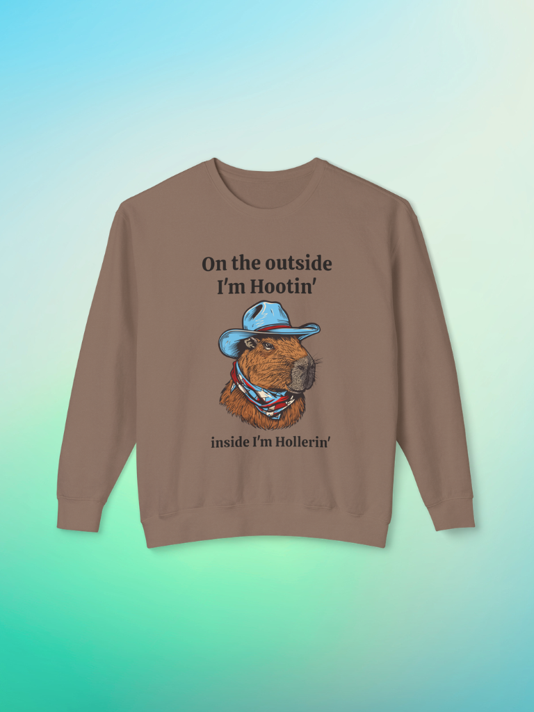 Brown crewneck sweatshirt featuring a colorful capybara wearing a blue cowboy hat and bandana, with text that reads ‘On the outside I’m Hootin’, inside I’m Hollerin’.’