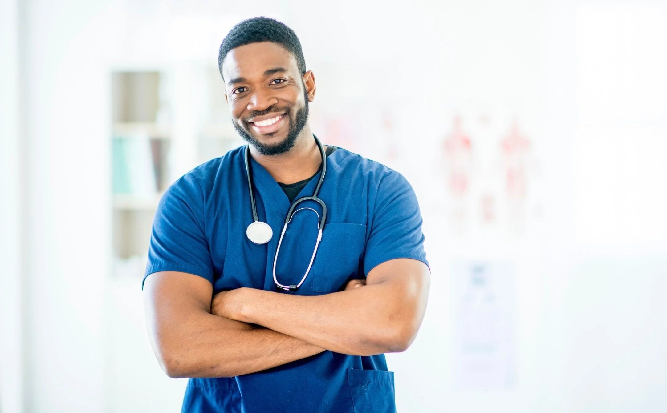 A smiling male healthcare worker wearing blue scrubs and a stethoscope around his neck, standing with arms crossed in a bright medical environment.