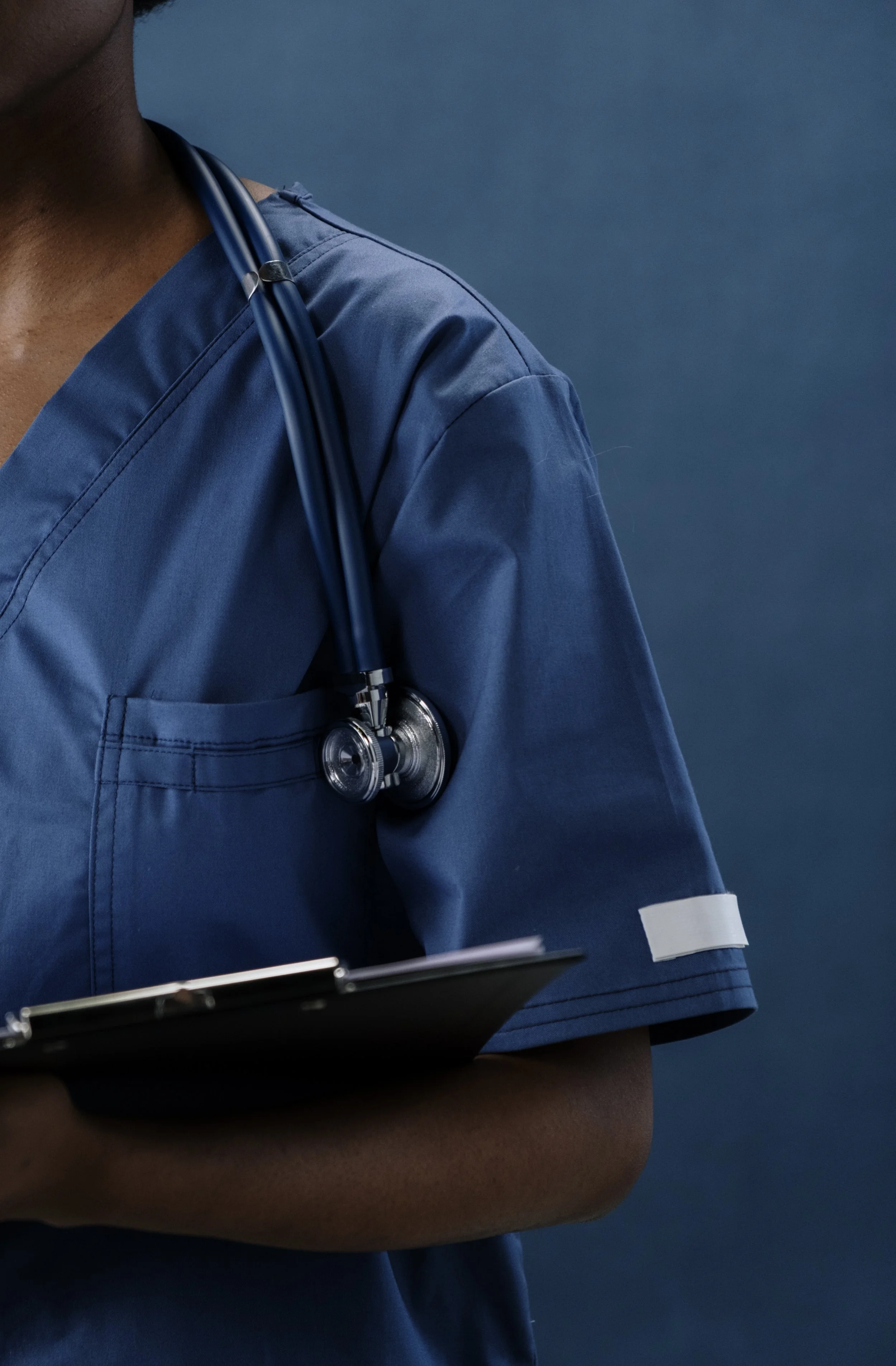 Close-up of a medical professional wearing blue scrubs with a stethoscope around their neck, holding a clipboard.