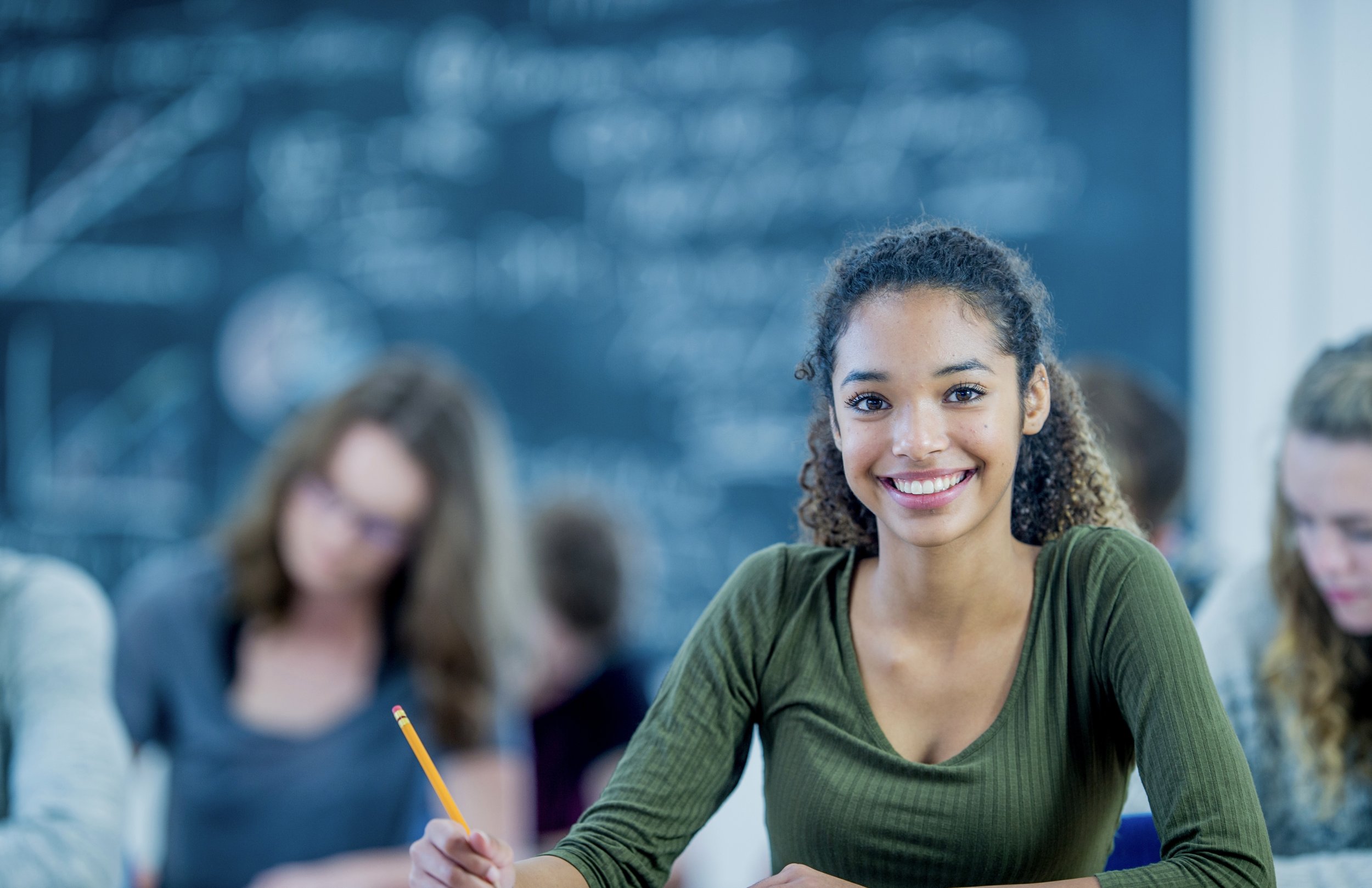 Smiling young woman in a classroom, with other students in the background, holding a pencil and engaging in learning.