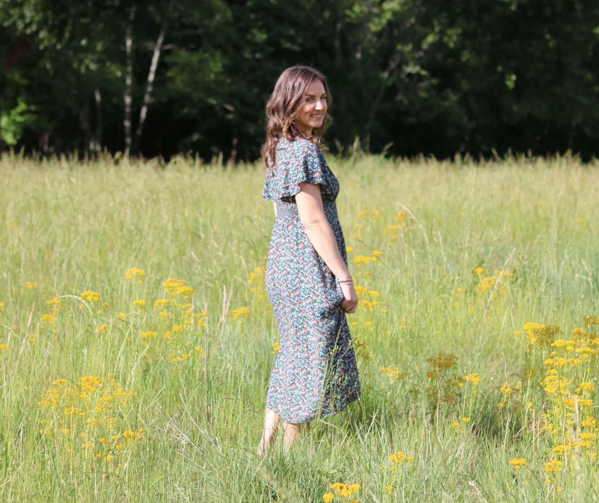 A woman standing in a grassy field with yellow flowers, wearing a floral dress and smiling, with trees in the background.