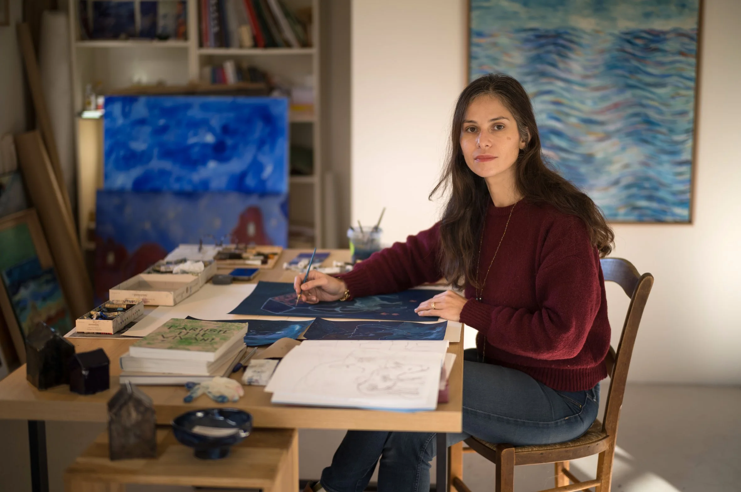 Jeanne-Sophie de Peretti assise et en train de peindre dans son atelier avec des peintures et des livres sur la table.