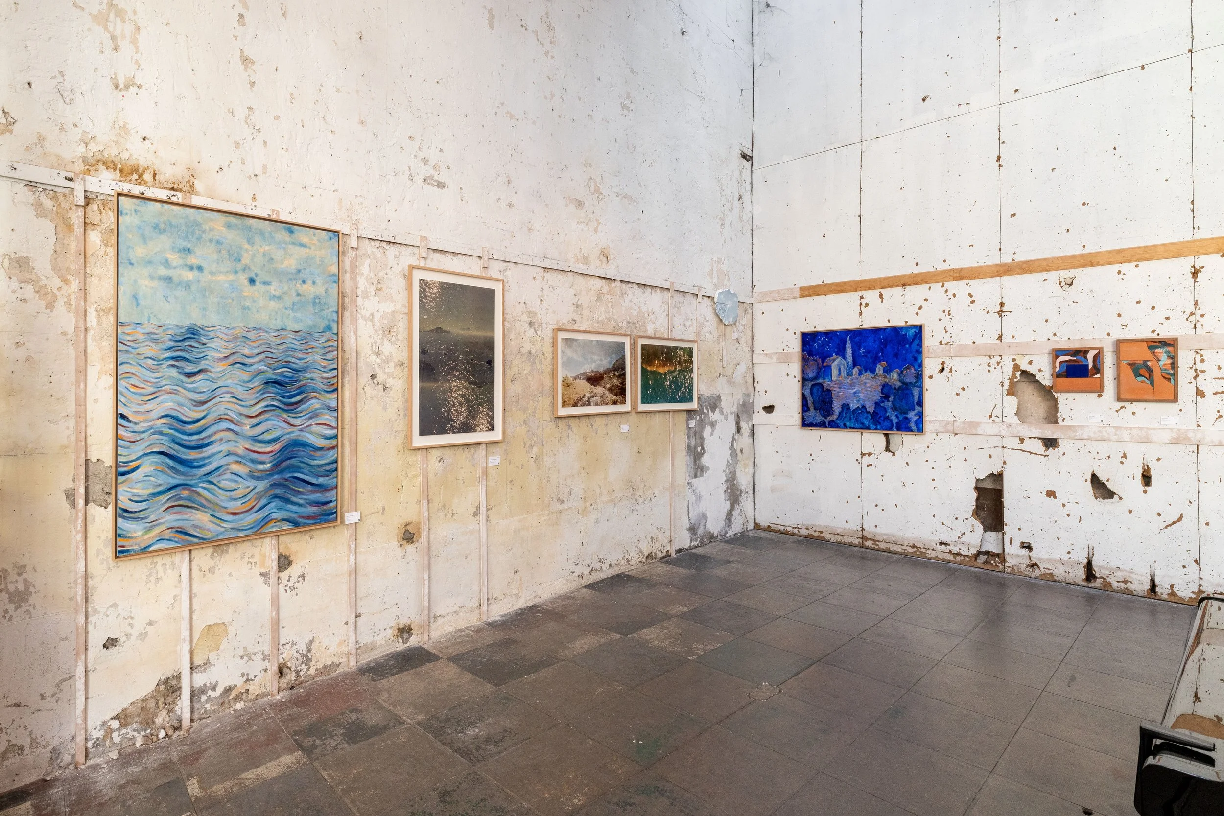 Galerie d'art avec plusieurs peintures suspendues sur un mur intérieur en mauvais état, murs cabossés et peints en blanc, sol en carrelage gris. Certaines œuvres sont des paysages ou des scènes marines. Exposition à l'Atelier Lardeur