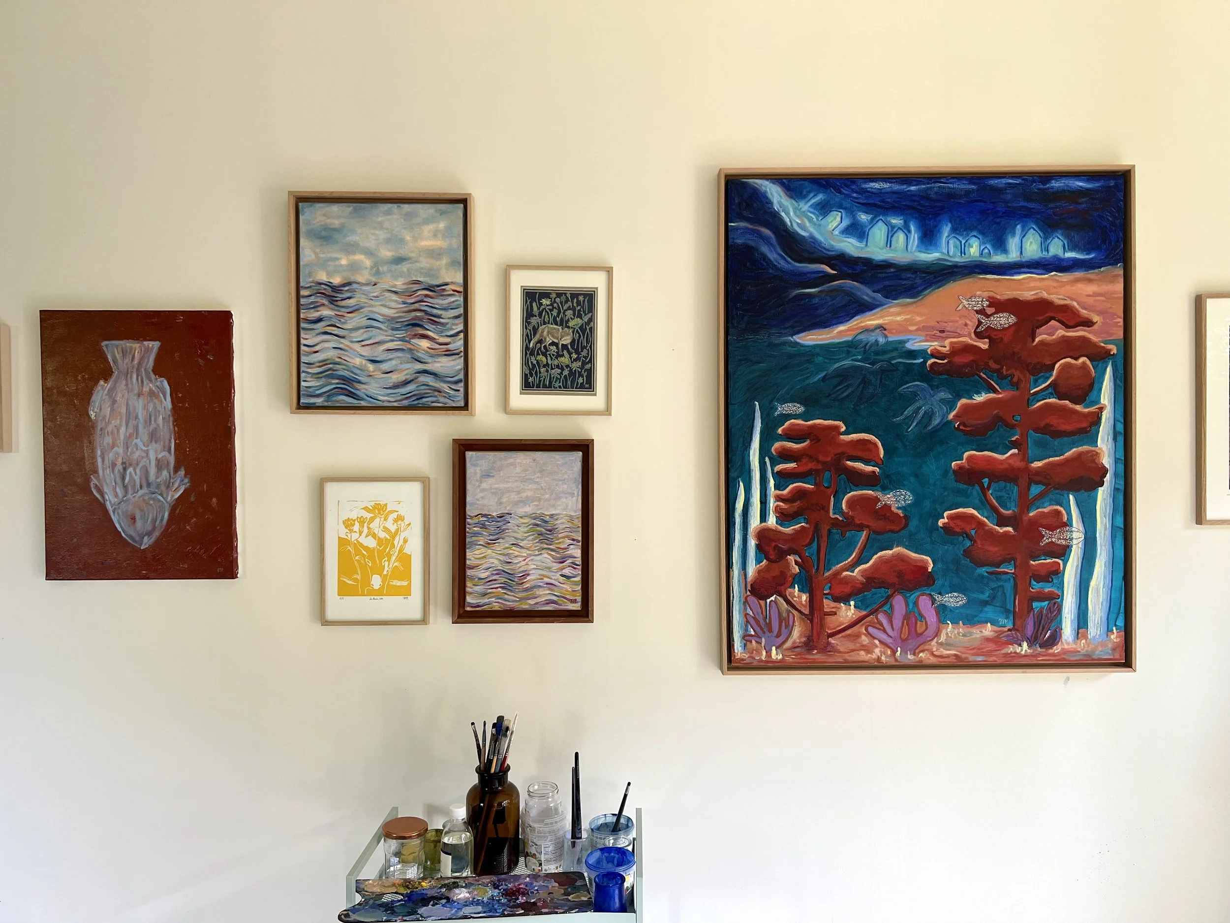 Dans l'atelier de Jeanne-Sophie de Peretti. Une collection de peintures accrochées à un mur, avec un meuble en dessous contenant des pinceaux, des pots de peinture et des verres.