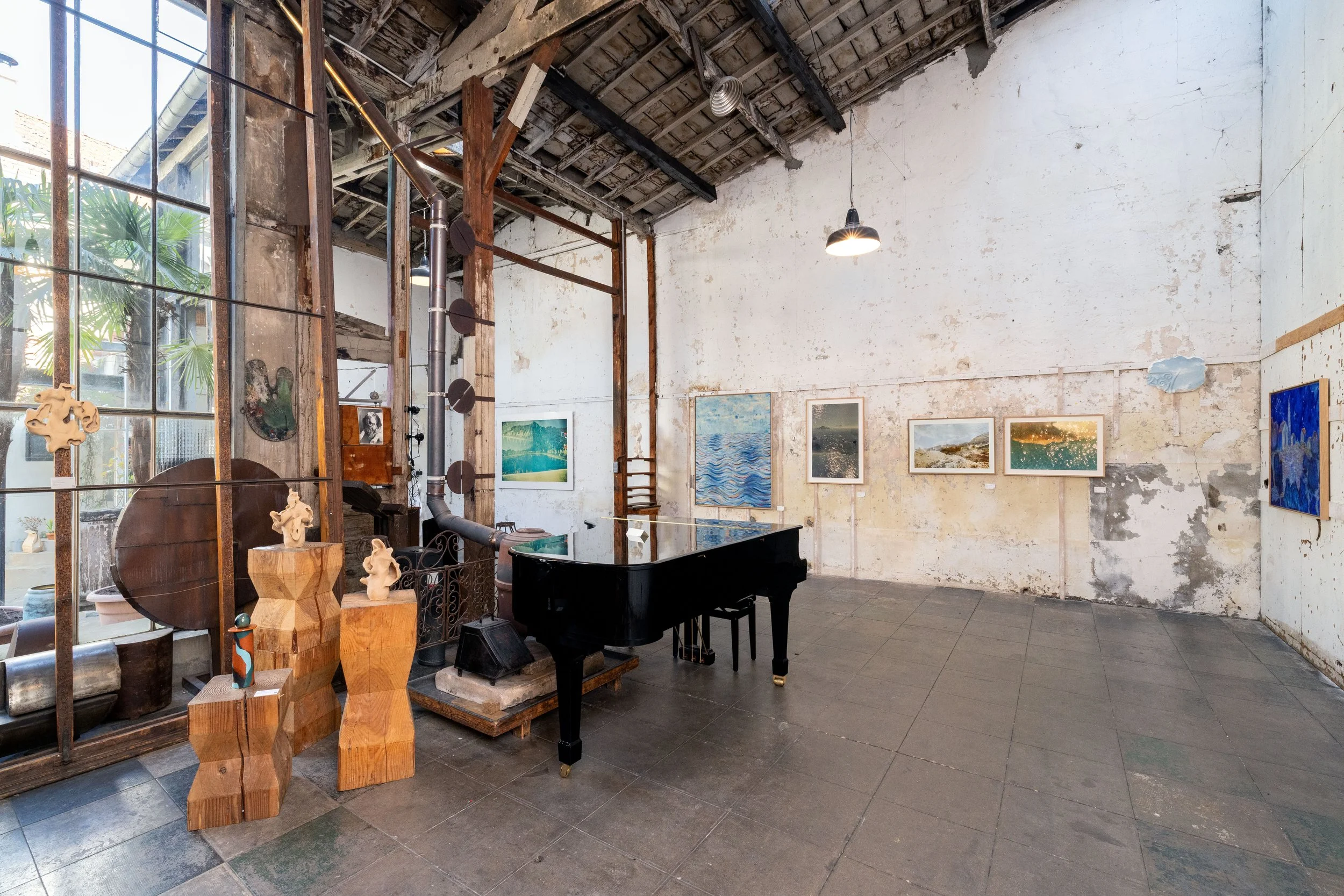 Une salle d'exposition d'art avec un piano noir, plusieurs œuvres d'art accrochées au mur, et des sculptures en bois sur des supports en bois. Le mur est ancien avec des murs en pierre ou en plâtre et un plafond en bois. C'est l'Atelier Lardeur.
