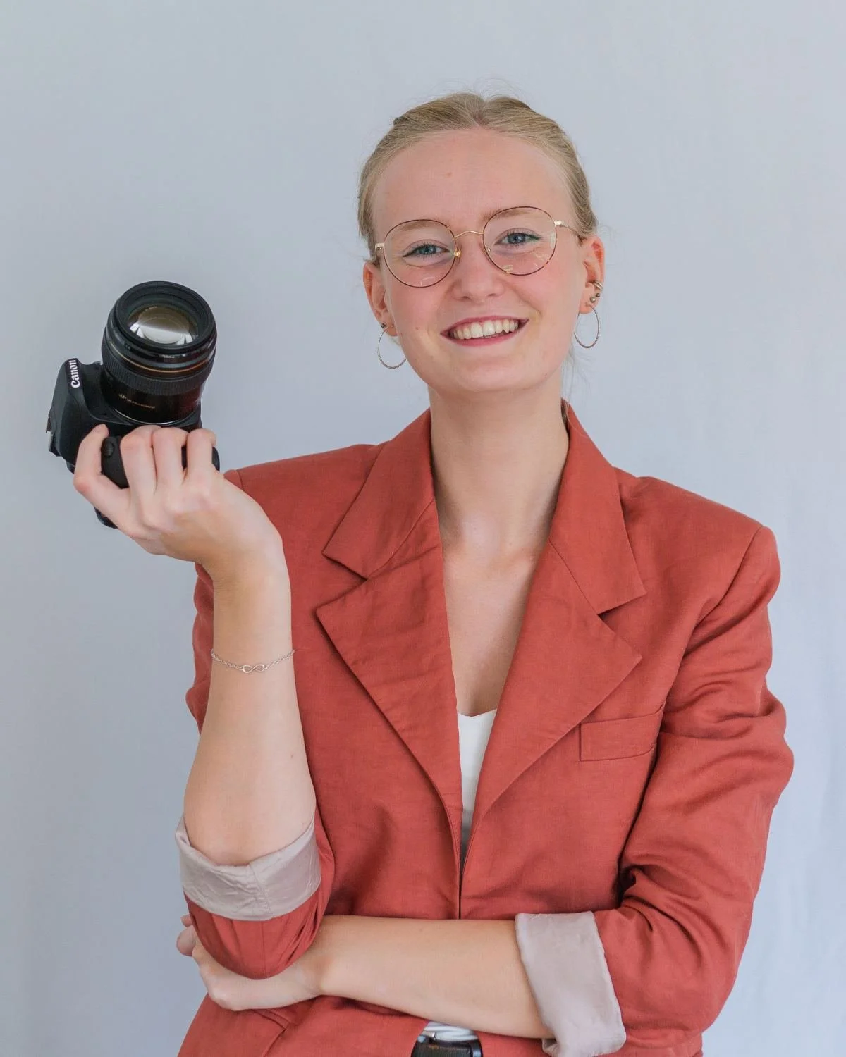 Frau lächelt in die Kamera. Sie trägt einen orangenen Blazer und in der rechten Hand eine Kamera.