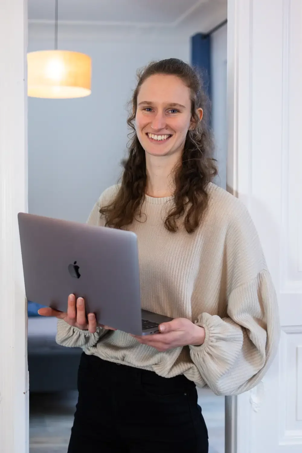 Webdesignerin Maike lehnt mit beigem Oberteil, schwarzer Hose und Laptop in Händen an weißer Flügeltür