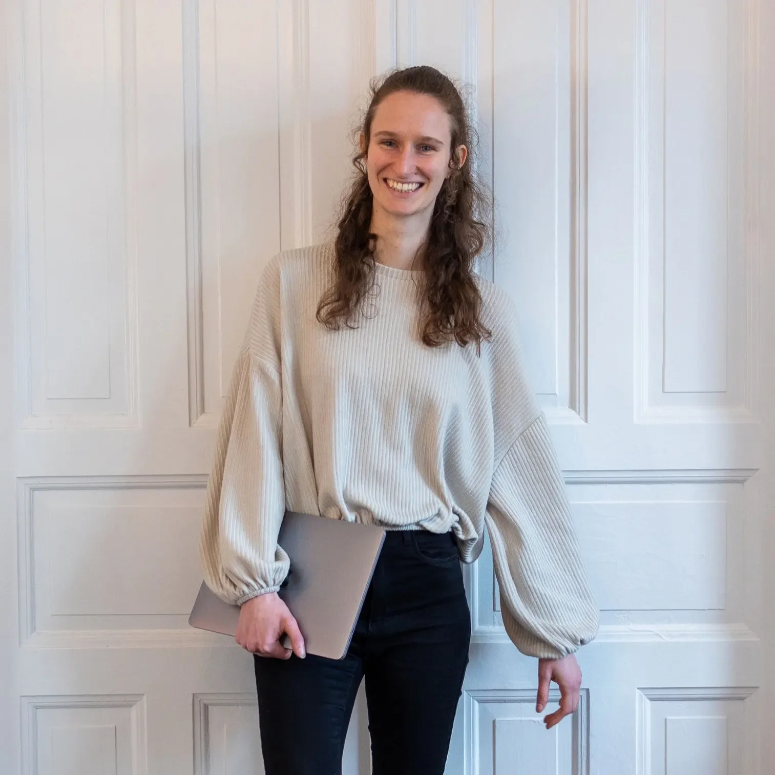 Maike im beigen Pullover vor weißer Altbautür, mit Laptop in der Hand