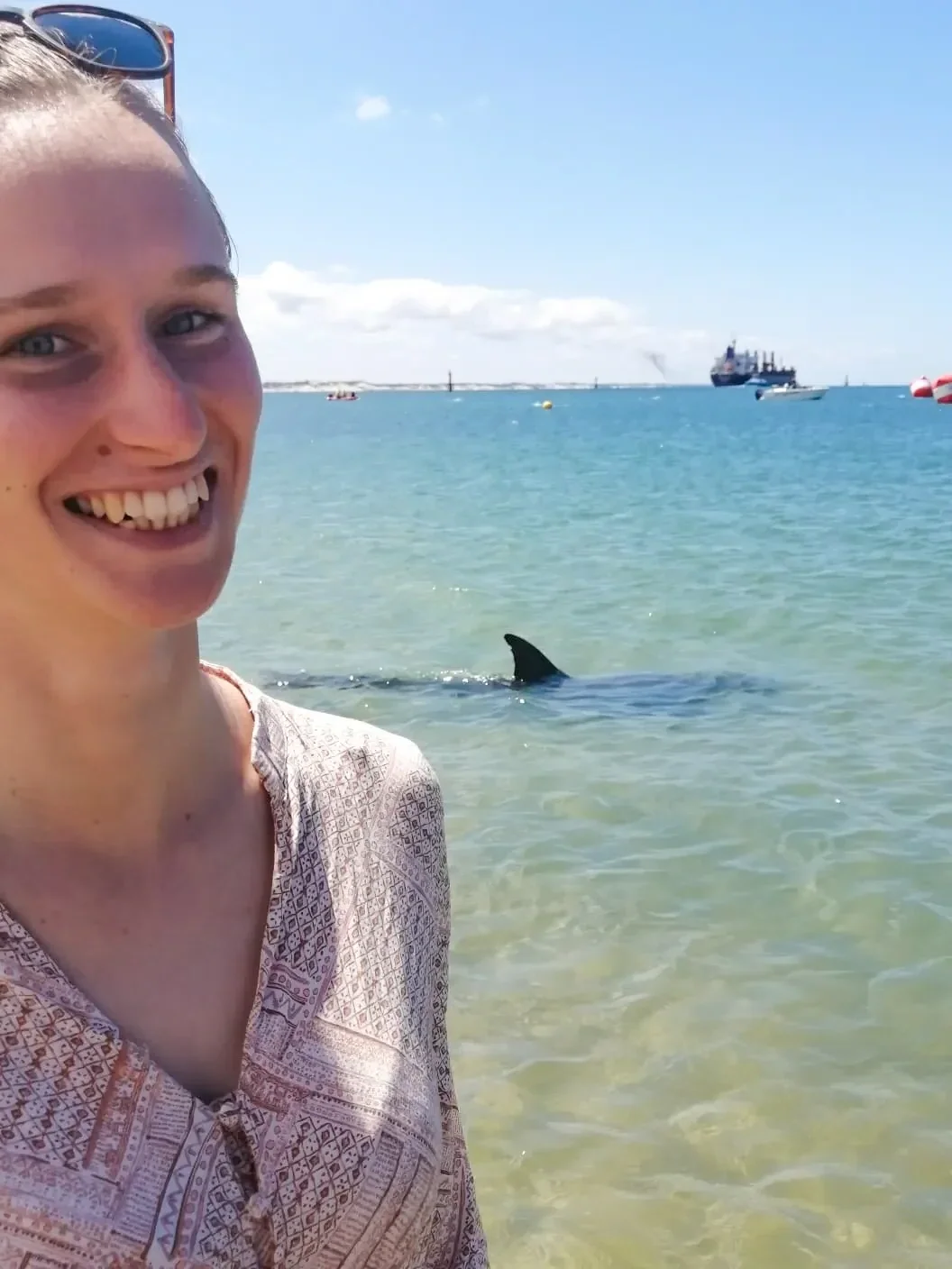 Maike mit Sonnenbrille am Meer, hinter ihr frei schwimmender Delfin