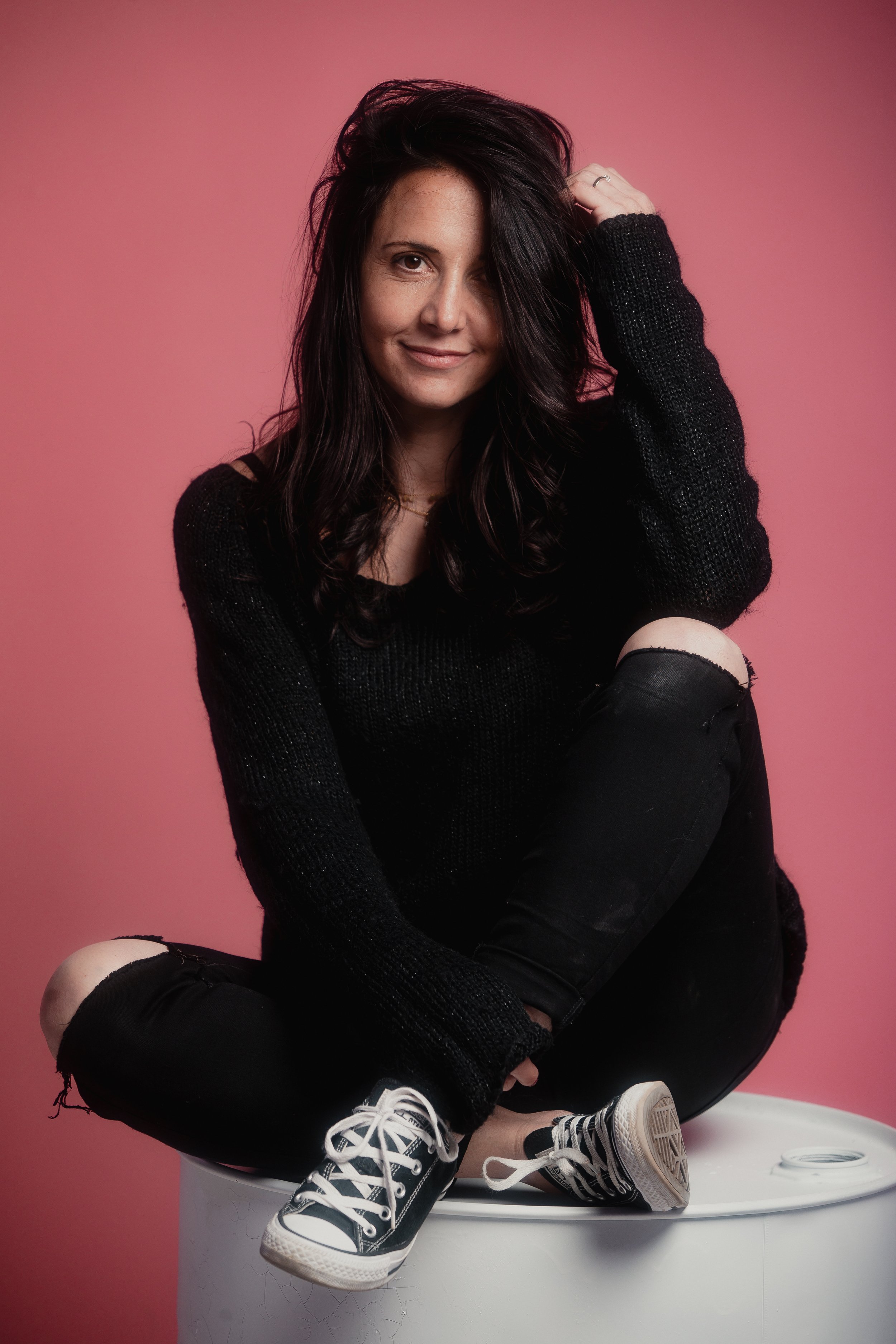 Jeune femme assise sur une surface blanche contre un fond rose, portant un pull noir, des jeans déchirés et des baskets Noirs et blancs, souriant.