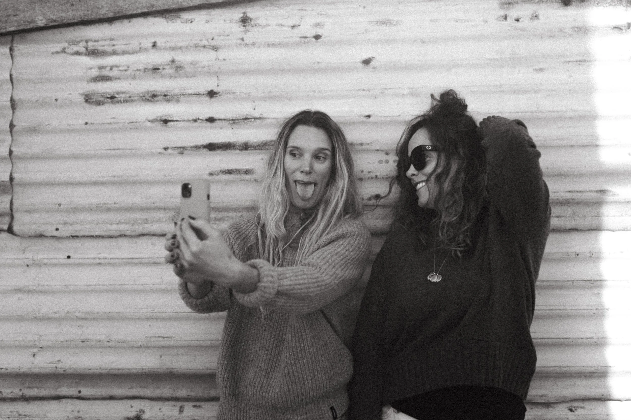 Two women wearing sunglasses, one with a striped shirt holding a magazine and the other talking on a vintage rotary phone, sitting at a marble table in a modern room.