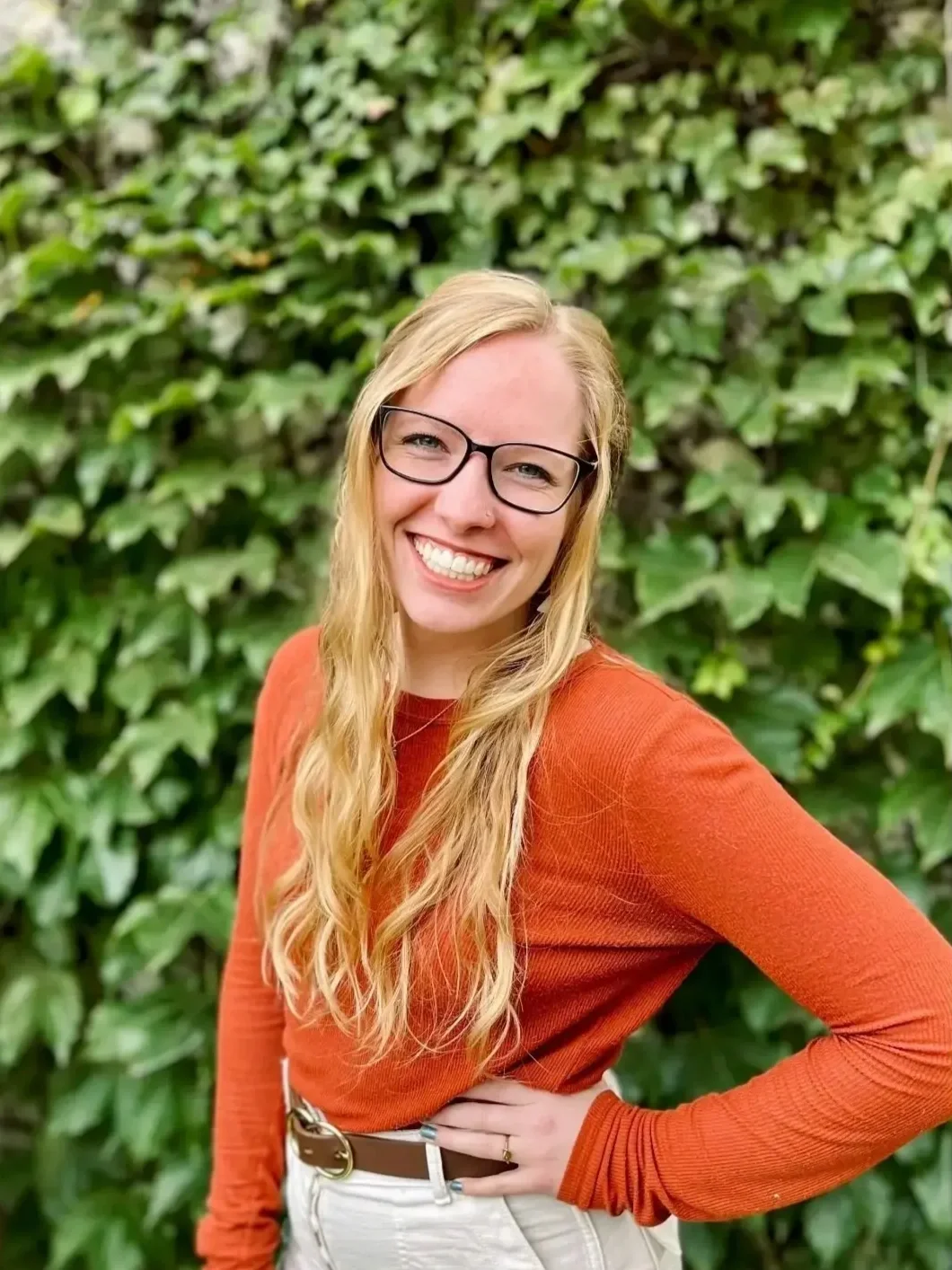 A smiling woman with long blonde hair, wearing glasses, an orange long-sleeve shirt, and white pants, standing in front of a lush green leafy background.