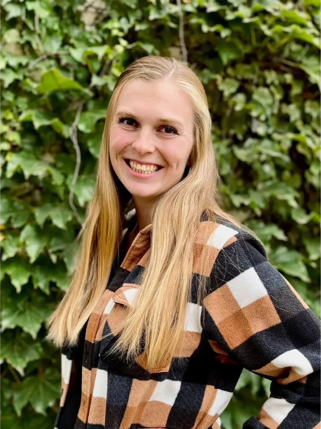 A young woman with long blonde hair smiling outdoors in front of green leafy foliage.
