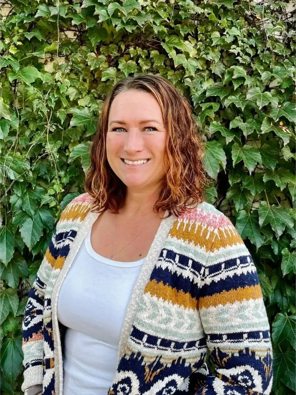 A woman with curly brown hair and blue eyes standing in front of a leafy green hedge, smiling at the camera, wearing a white shirt and a colorful patterned cardigan.