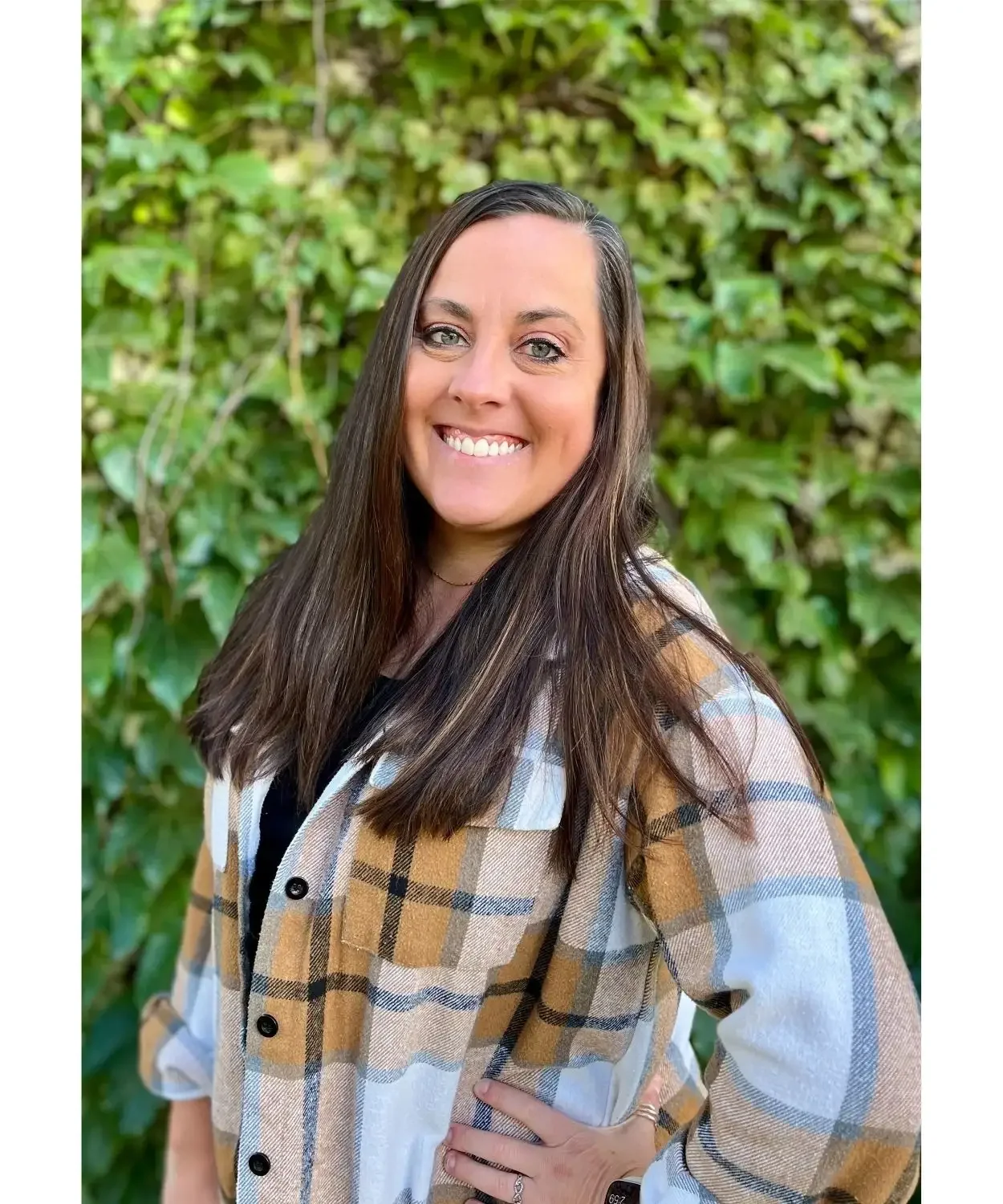 A woman with long dark hair, wearing a beige, white, and black plaid shirt, smiling outdoors in front of green leafy plants.