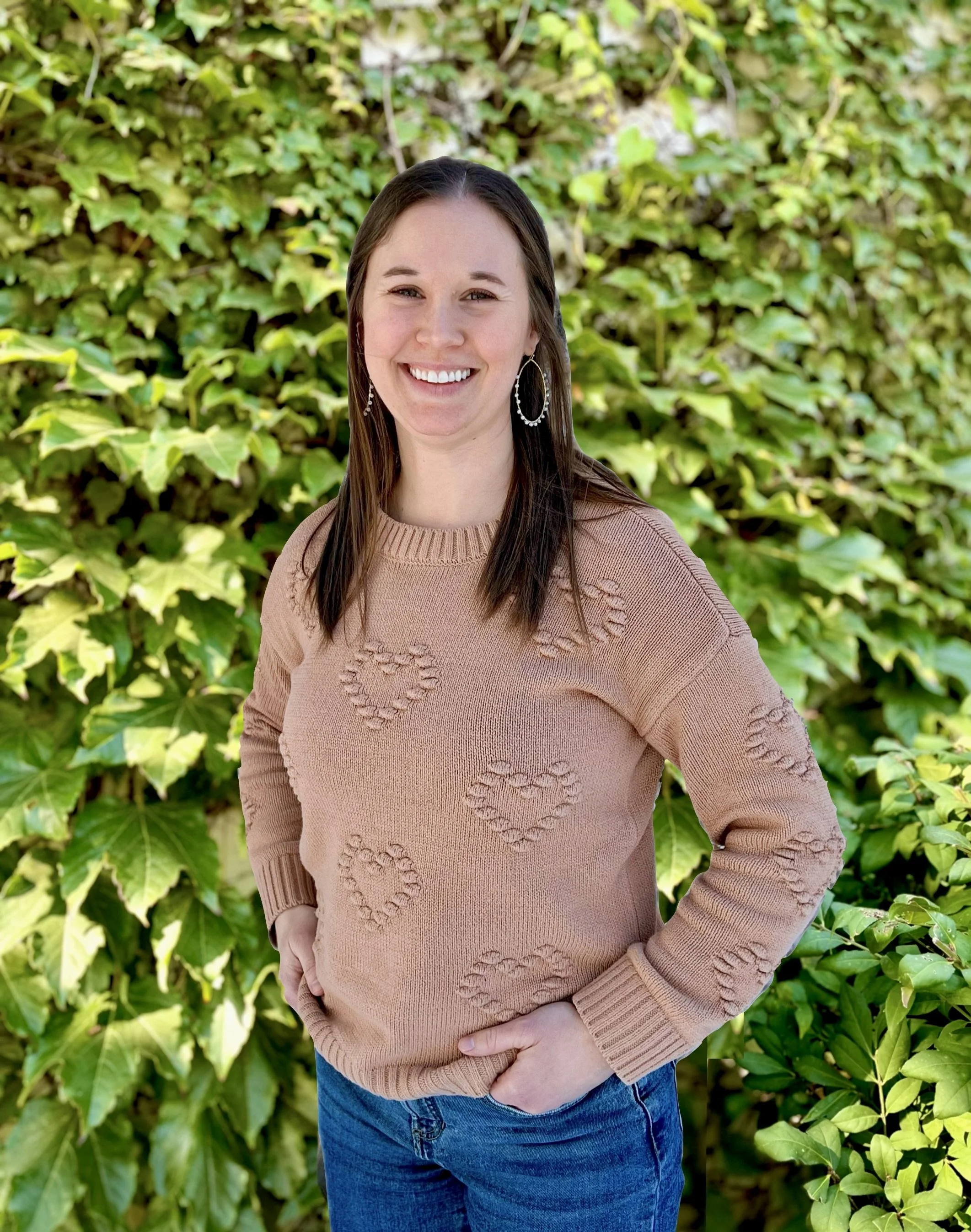 A young woman with shoulder-length brown hair, smiling and wearing a black top, standing outdoors in front of lush green foliage.