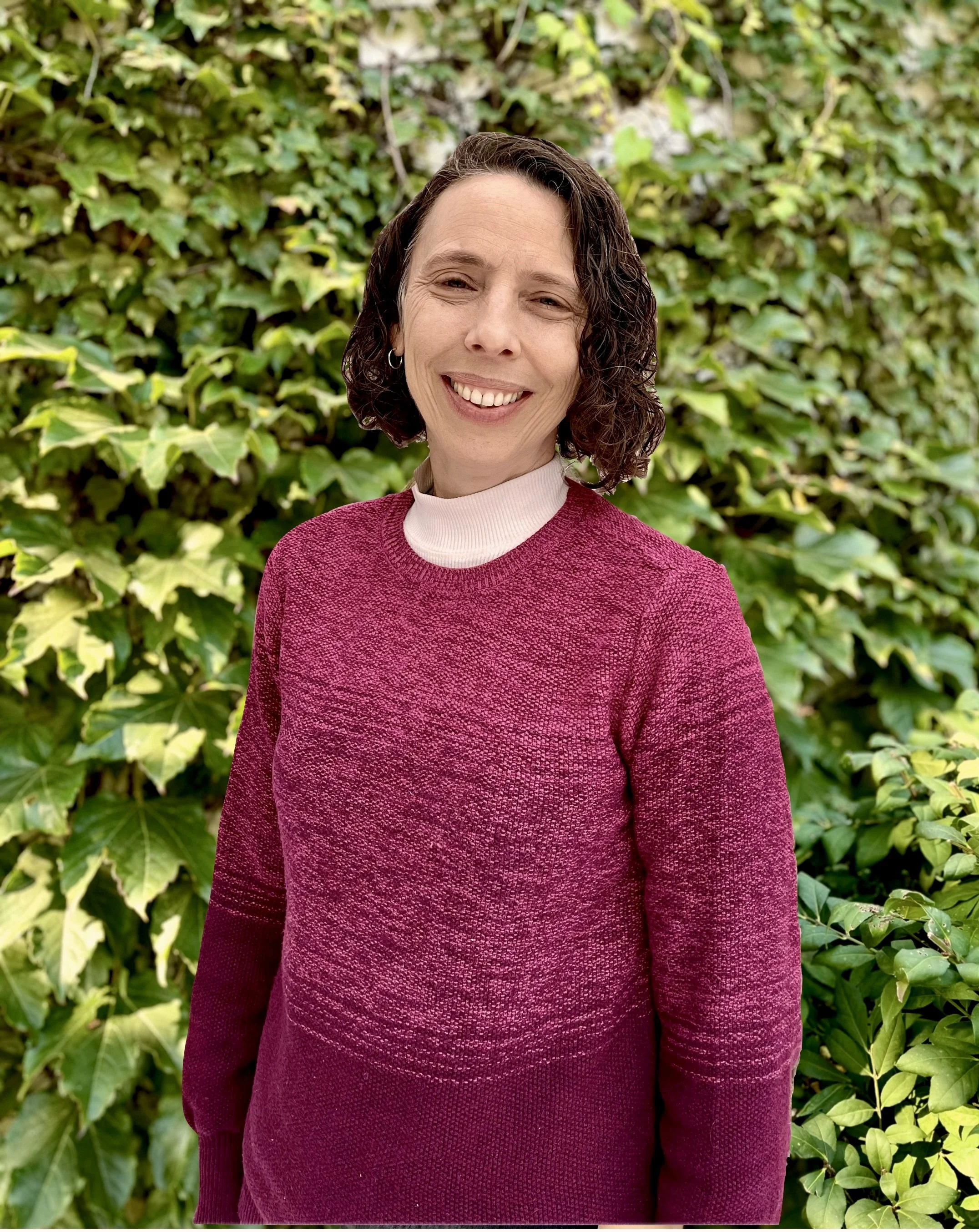 A woman with curly brown hair smiling, taken indoors with part of a window and a wooden chair visible in the background.