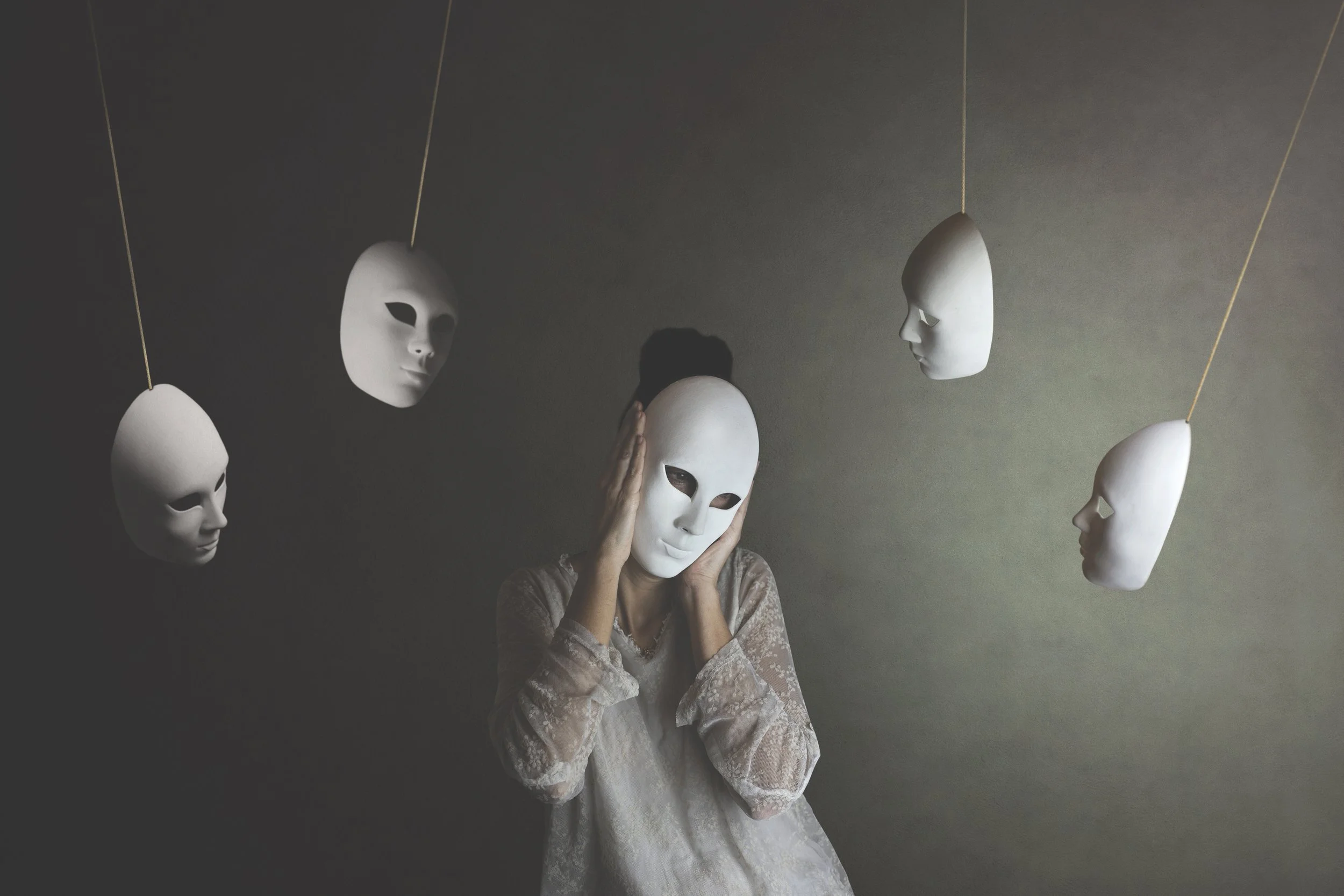 Person wearing a white mask surrounded by four hanging white masks on dark background.