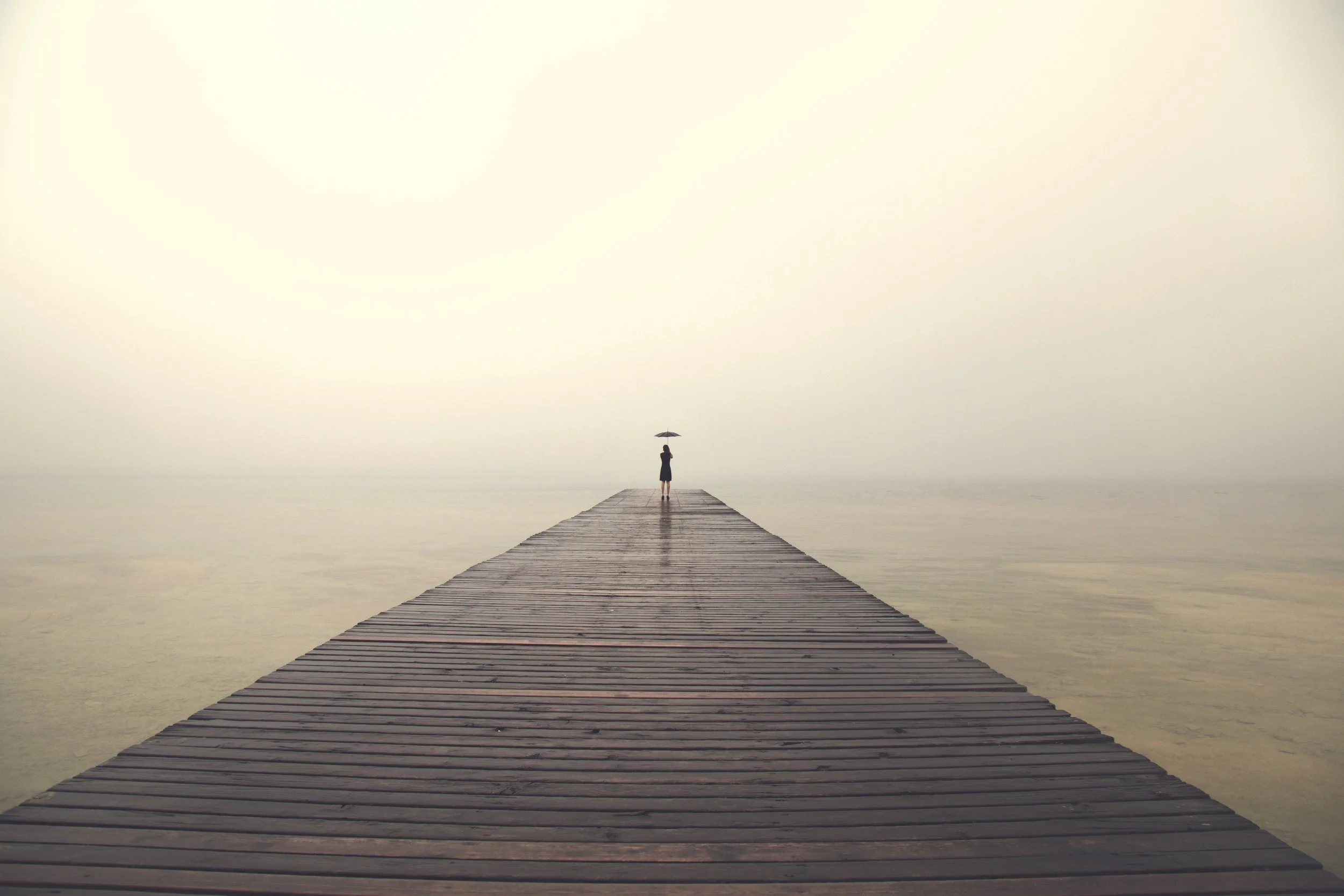 Person holding umbrella on wooden pier in foggy landscape
