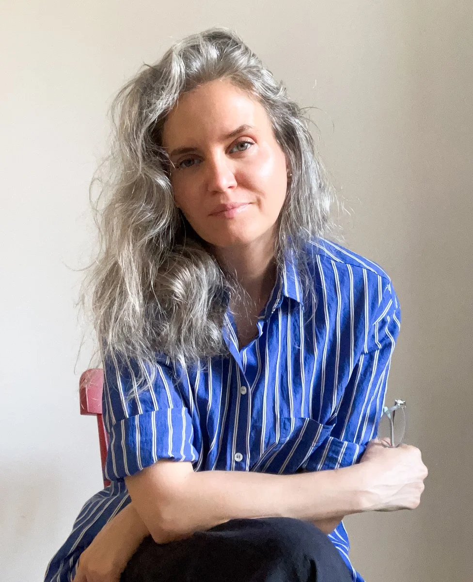 A woman with shoulder-length gray curly hair wearing a blue and white striped shirt sitting against a plain beige wall.