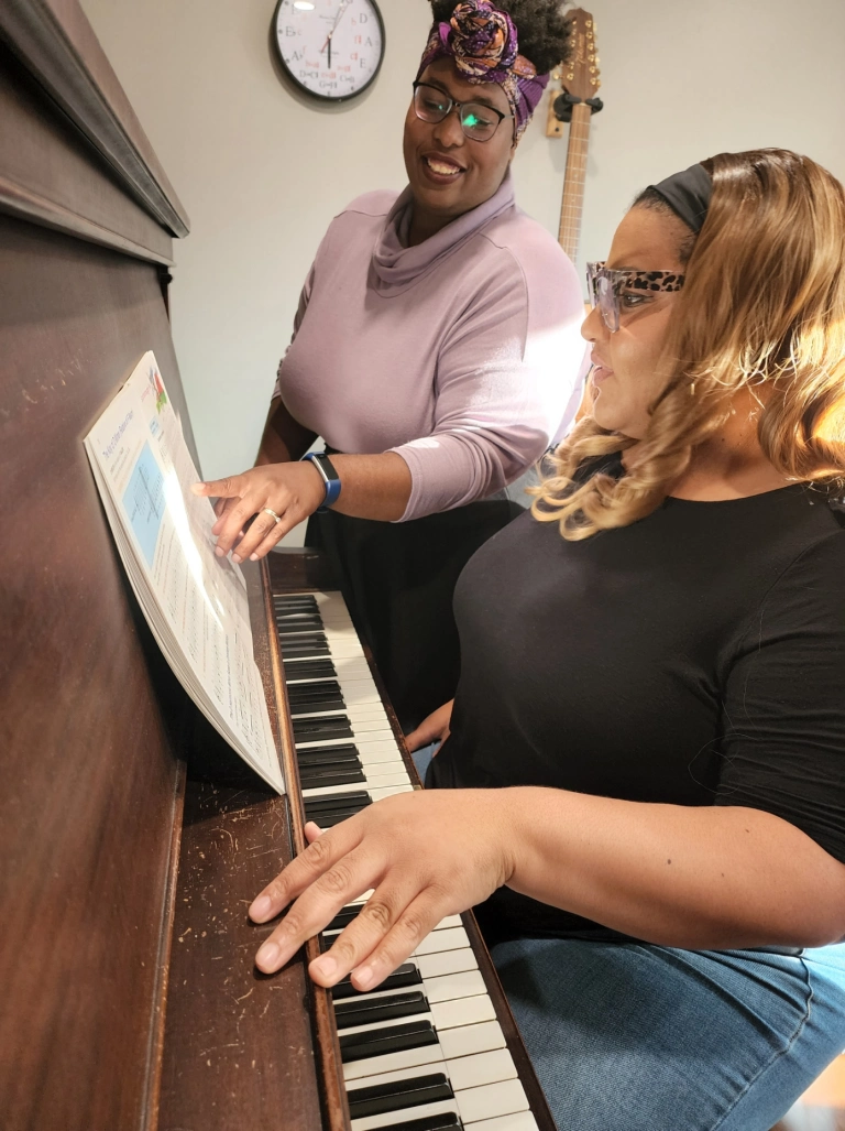 Two women at a piano. One woman with curly hair and glasses is standing, pointing at music sheet, and smiling, while the other woman with blonde hair and glasses is sitting, playing the piano. A clock is on the wall behind them.