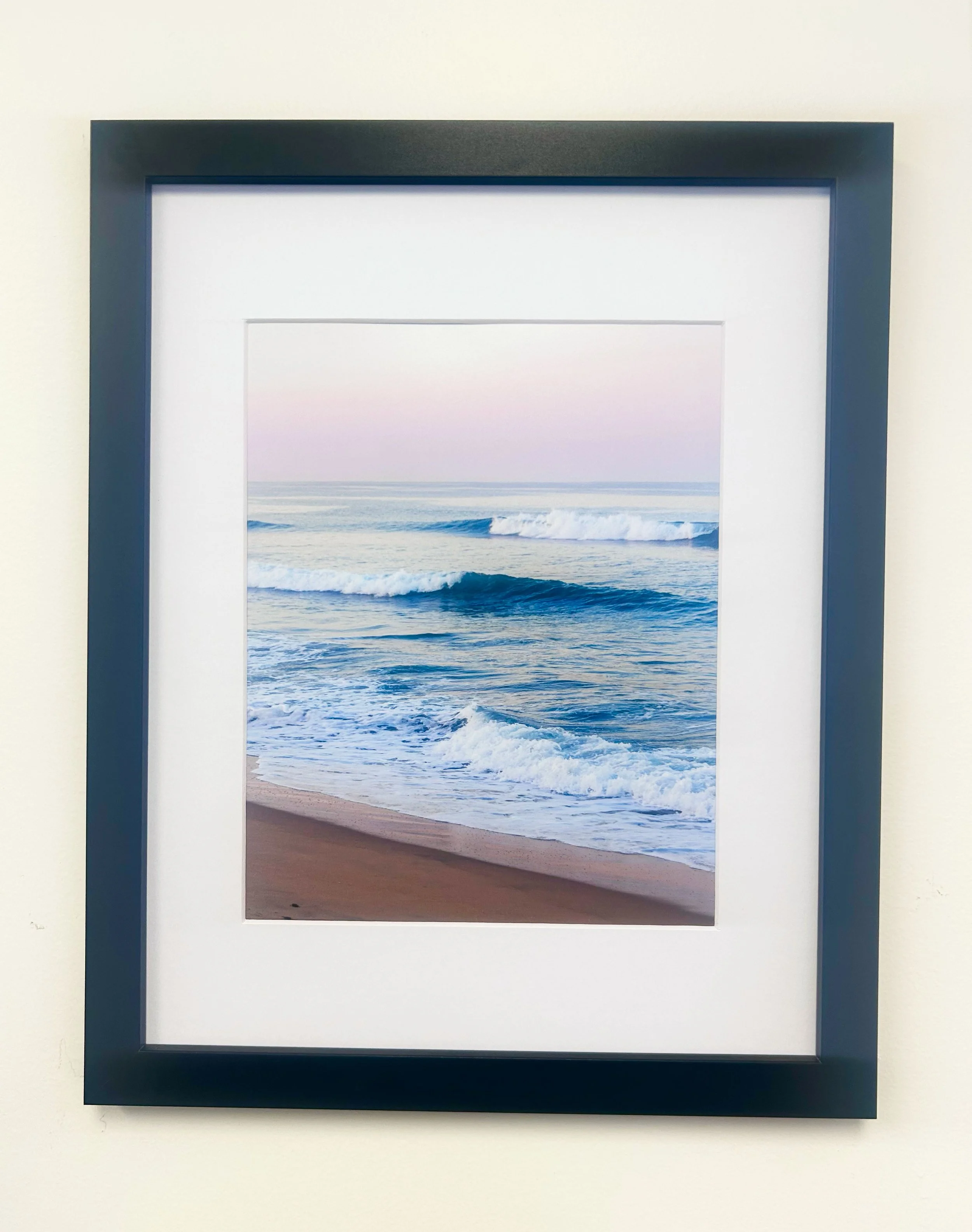A framed photograph of the ocean with waves crashing on the beach, viewed through a black picture frame with a white border.