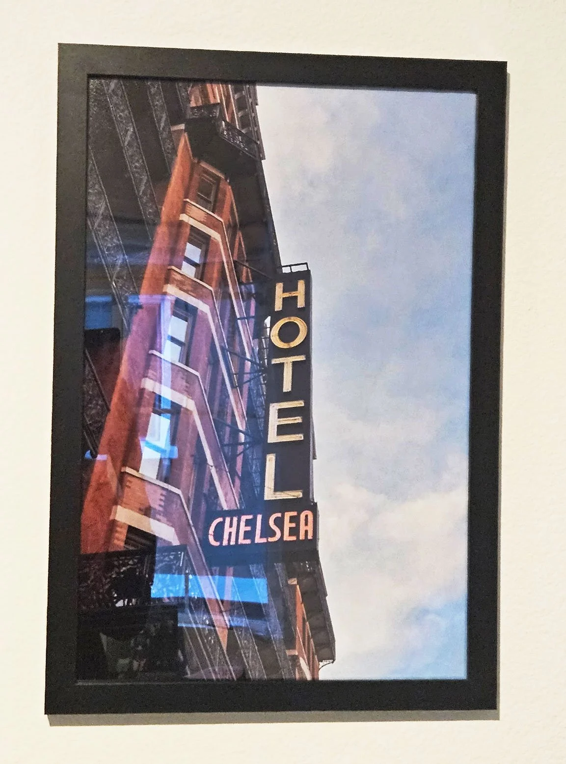 A photograph of a building with a sign that reads 'HOTEL CHELSEA'. The building has several windows and fire escapes, and the sky in the background is partly cloudy.