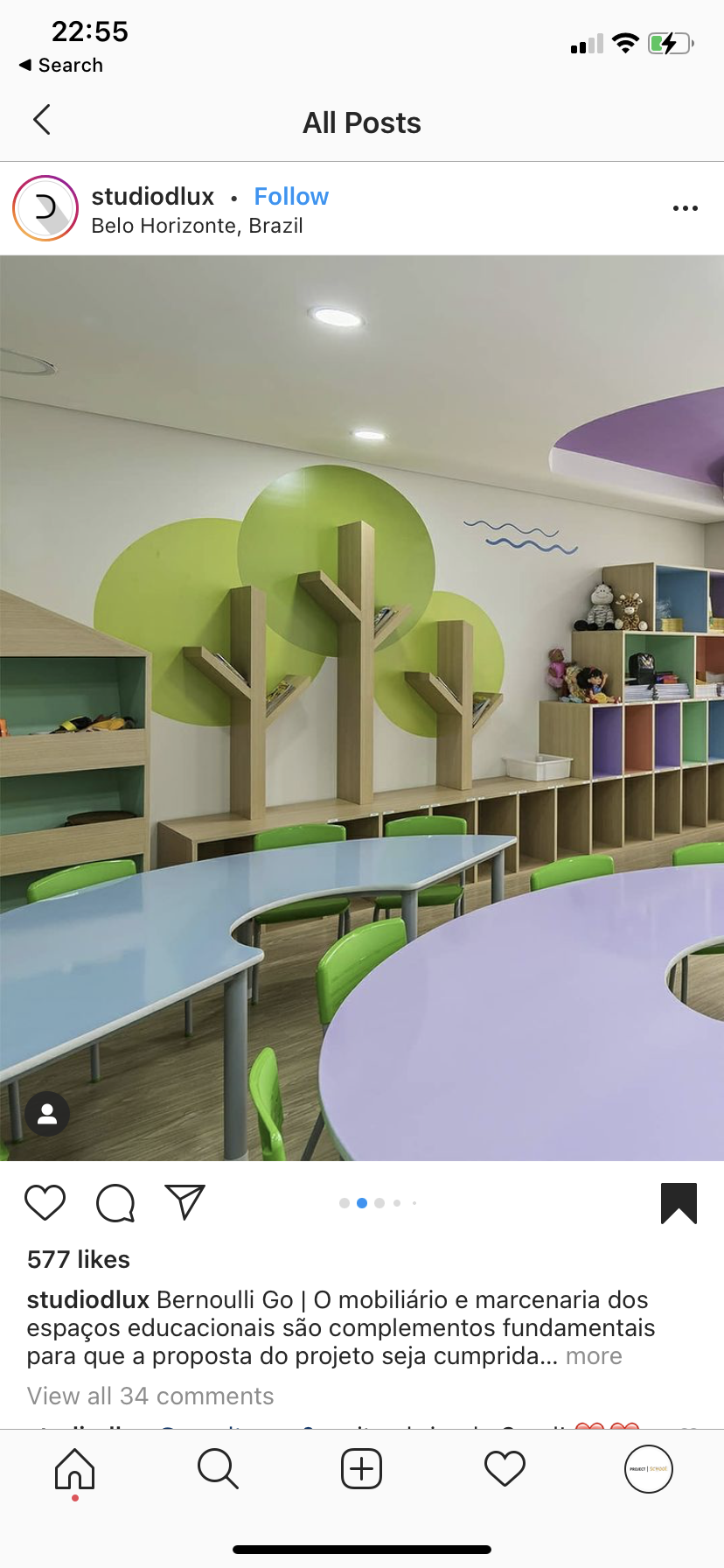 Colorful educational room with green, purple, and blue tables, green chairs, and decorative tree-shaped shelves on the wall filled with toys and books.