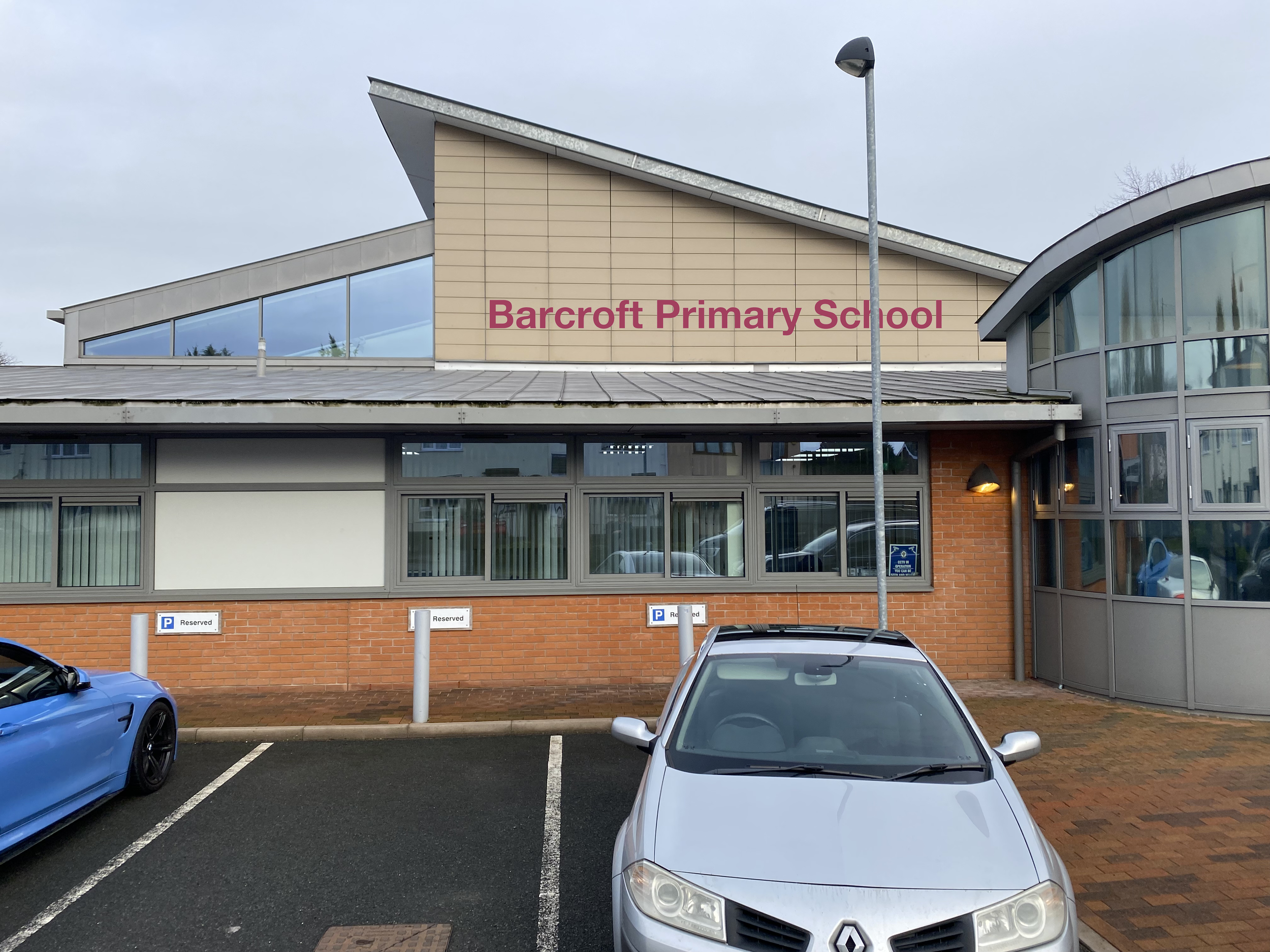 Front view of Barcroff Primary School building with parking lot showing reserved parking spots and cars, including a silver Renault and a blue sports car, on a cloudy day.