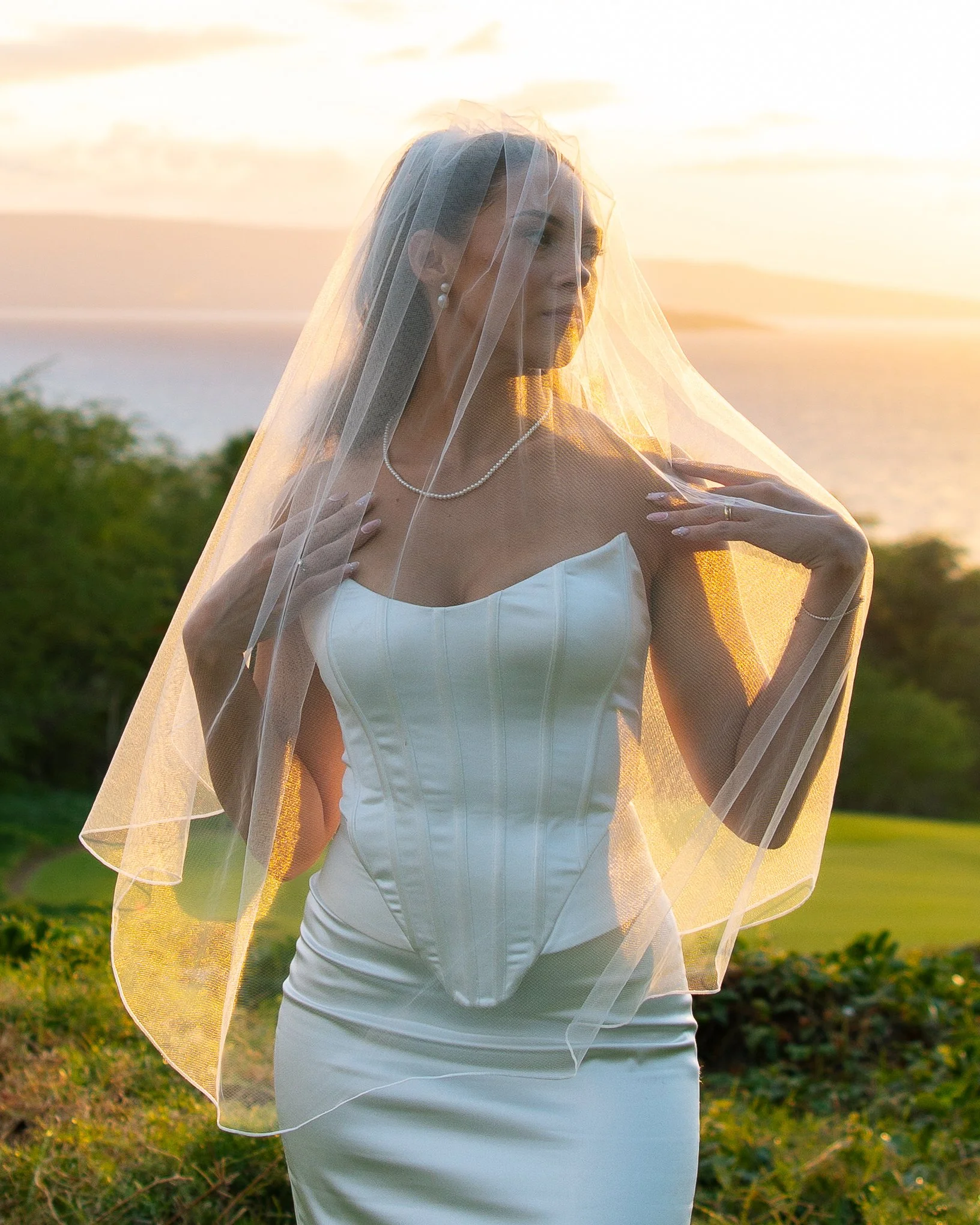 A bride in a white wedding dress and pearl necklace standing outdoors during sunset, wearing a veil, with greenery and water in the background.