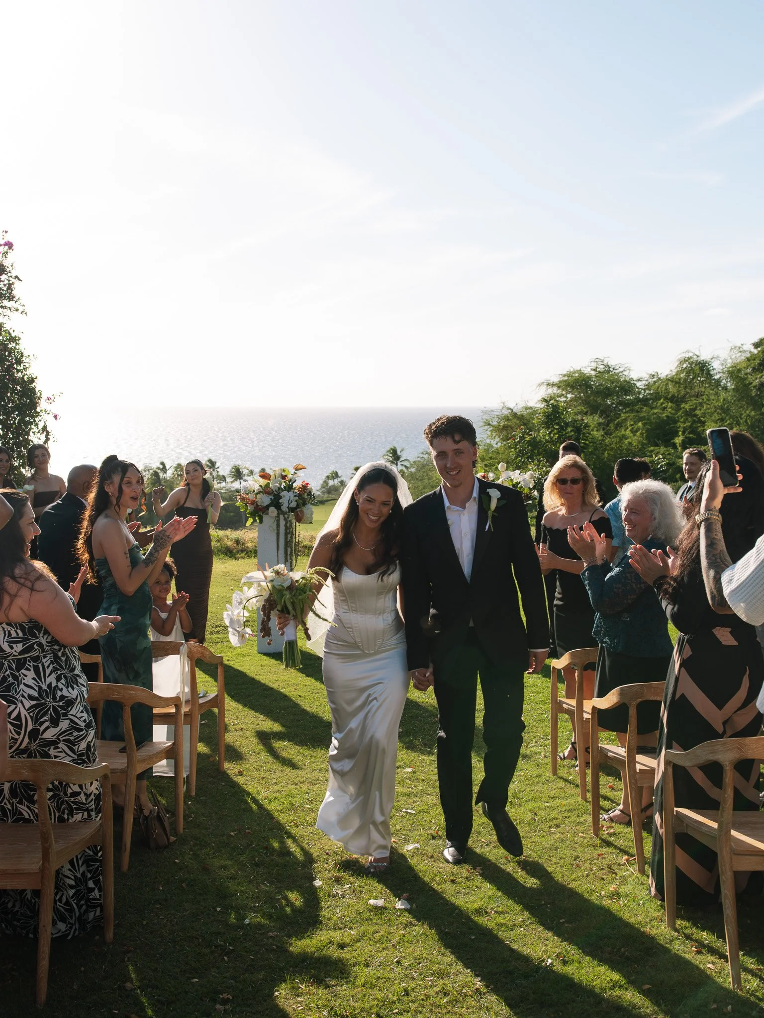 A bride and groom walking down the aisle hand in hand at an outdoor wedding ceremony, surrounded by guests clapping and celebrating. The background features a scenic view of the ocean and a bright, clear sky.