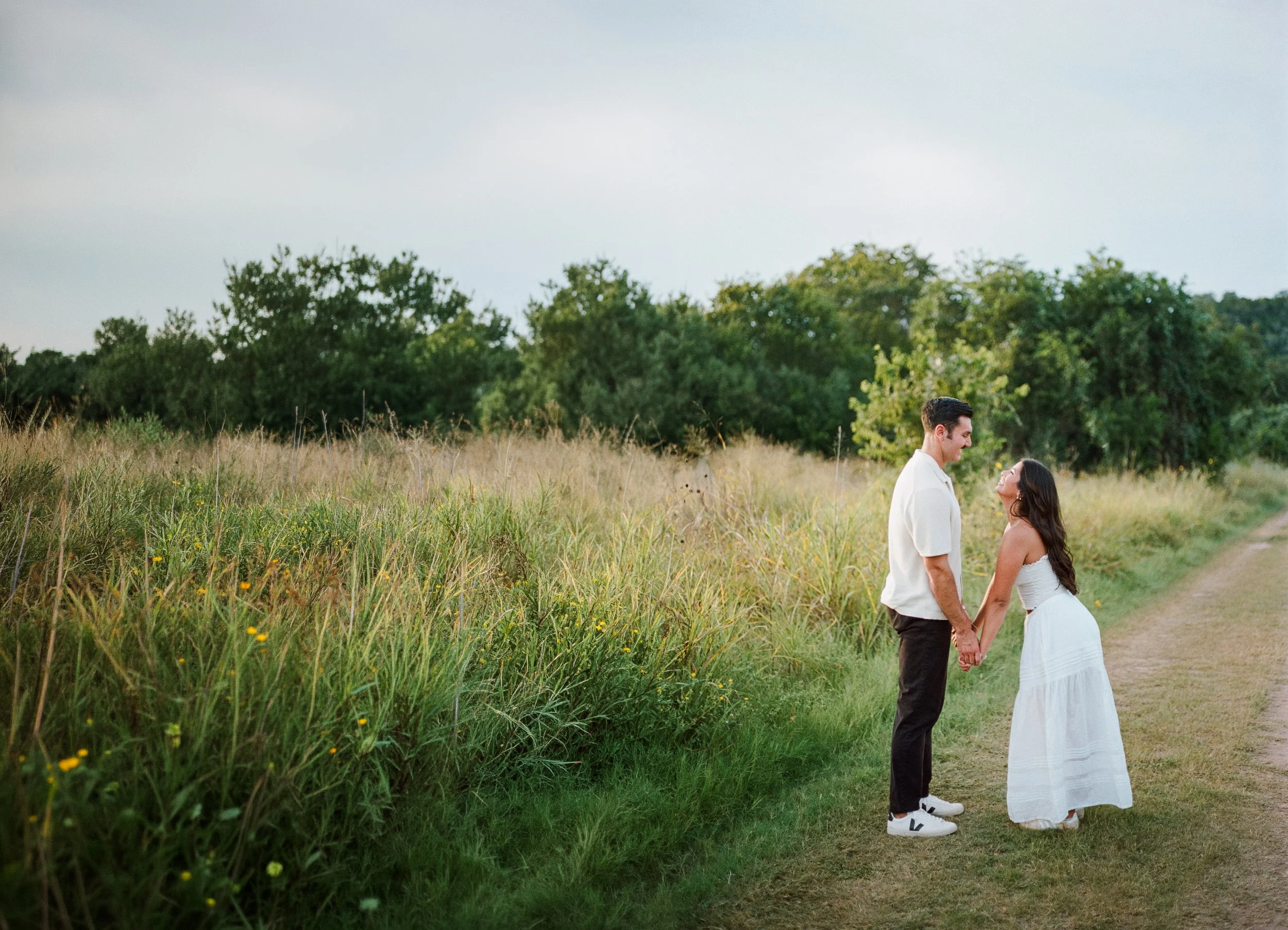A couple holding hands and smiling at each other standing in a field with tall grass and trees in the background on a cloudy day.