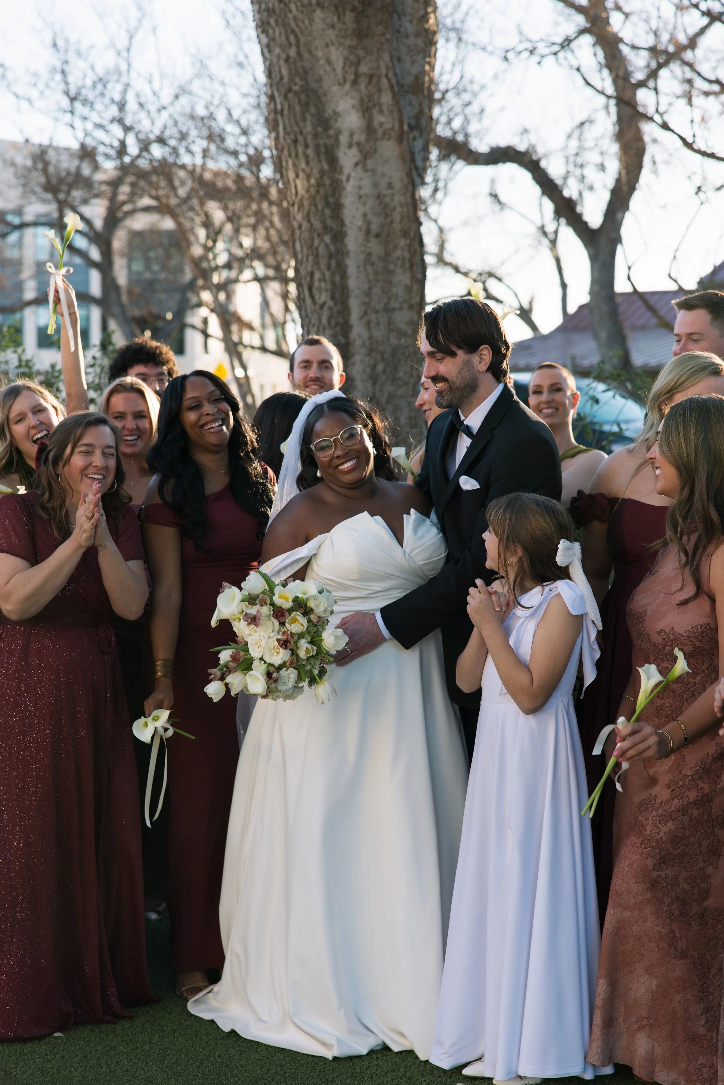 A group of people at a wedding, centered around a bride in a white dress and a groom in a black tuxedo, surrounded by bridesmaids and guests outdoors with trees and buildings in the background.
