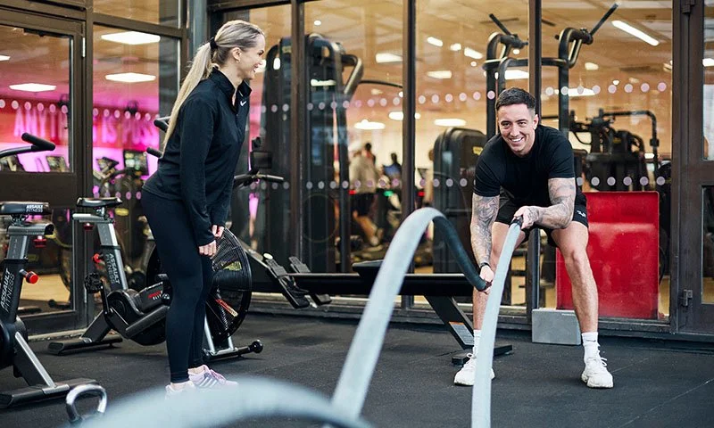 Personal trainer encouraging a client as he performs a battle ropes exercise on a commercial gym floor.