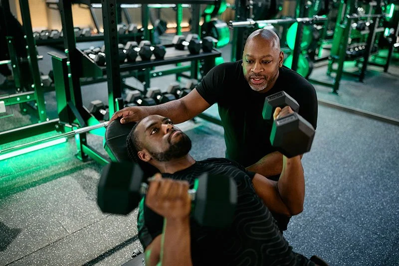 Self-employed personal trainer coaching a client through a dumbbell chest press in a UK commercial gym