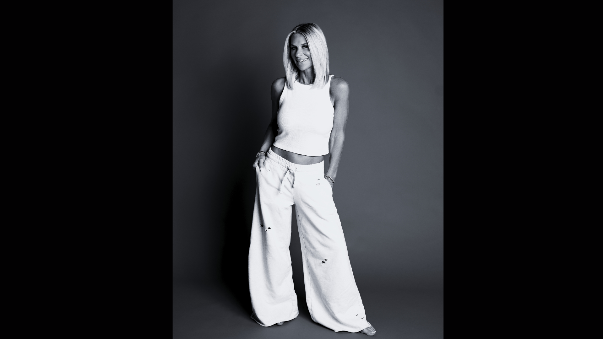 A woman smiling and posing with her hands in her pockets. She has shoulder-length blonde hair and is wearing a sleeveless white top and wide-leg distressed white pants, standing against a plain background.