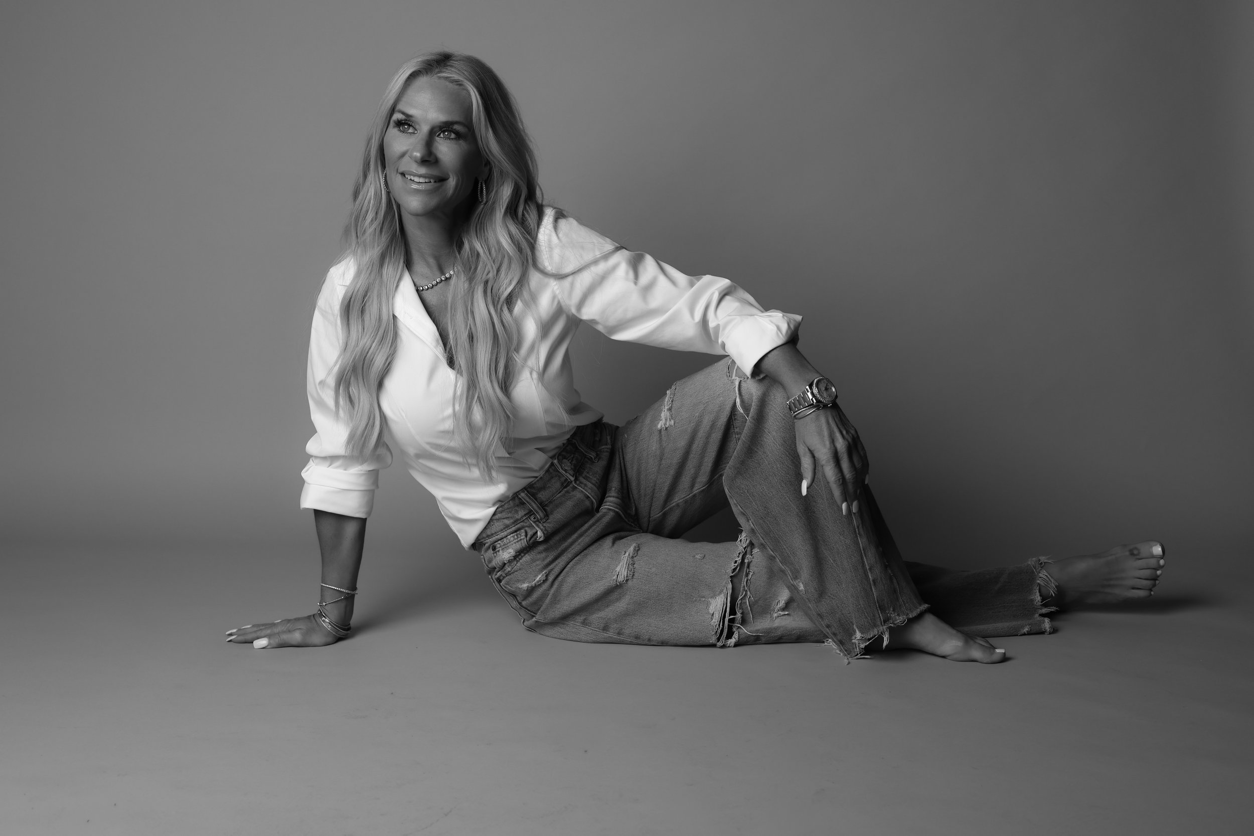 Black and white photo of a woman with long wavy hair, sitting on the floor with one arm supporting her, wearing a satin blouse, ripped denim jeans, jewelry, and a watch, against a plain background.