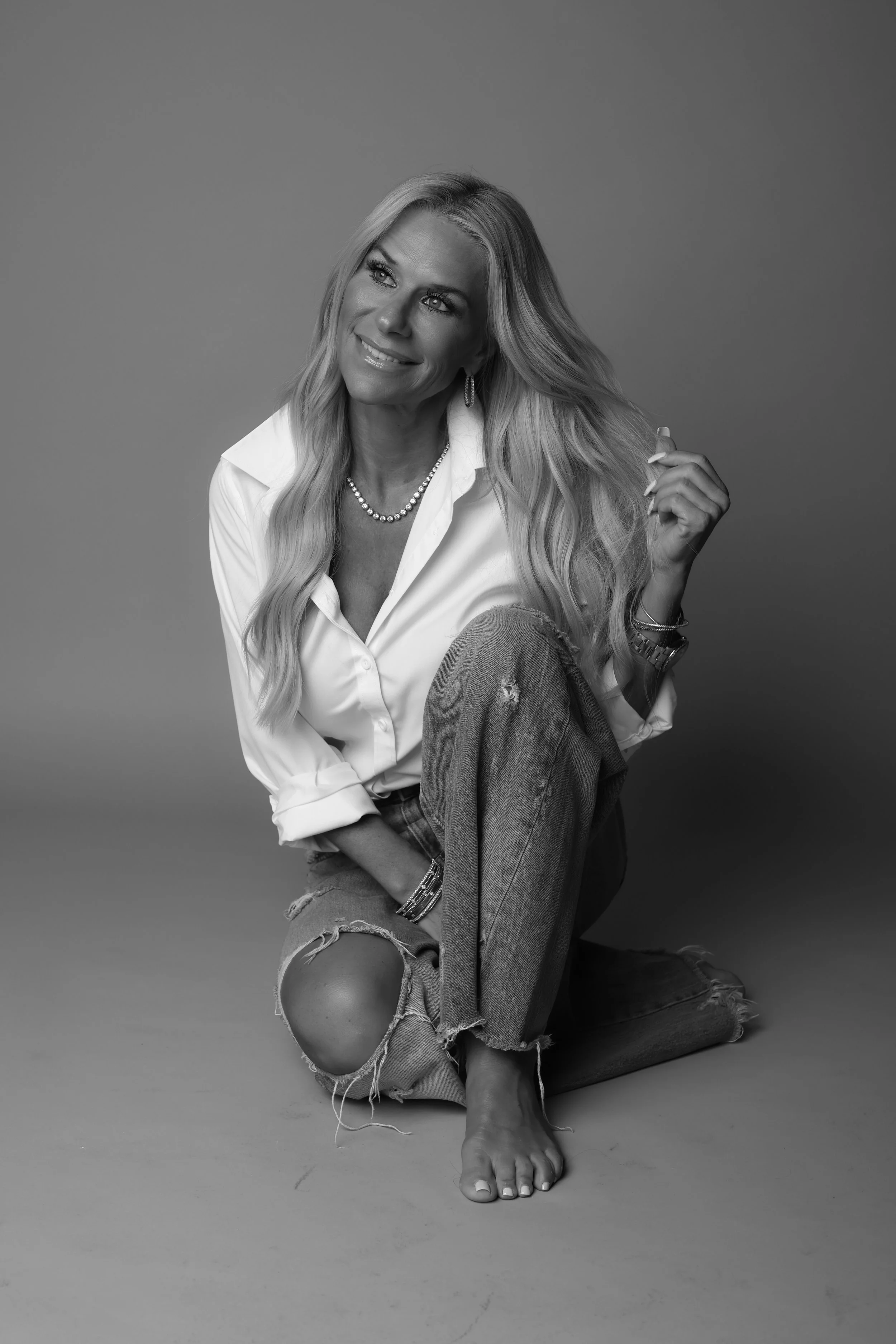 Black and white portrait of a smiling blonde woman with long hair, wearing a white shirt, jeans with a large knee hole, and accessories, sitting on the floor against a plain background.