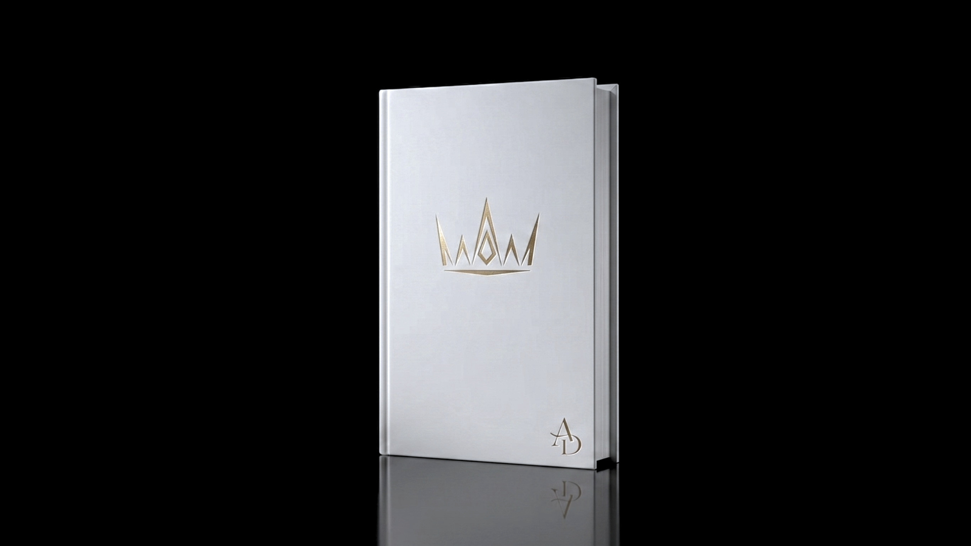 A white journal with a gold crown design and the initials 'AD' on the cover, standing upright against a black background.