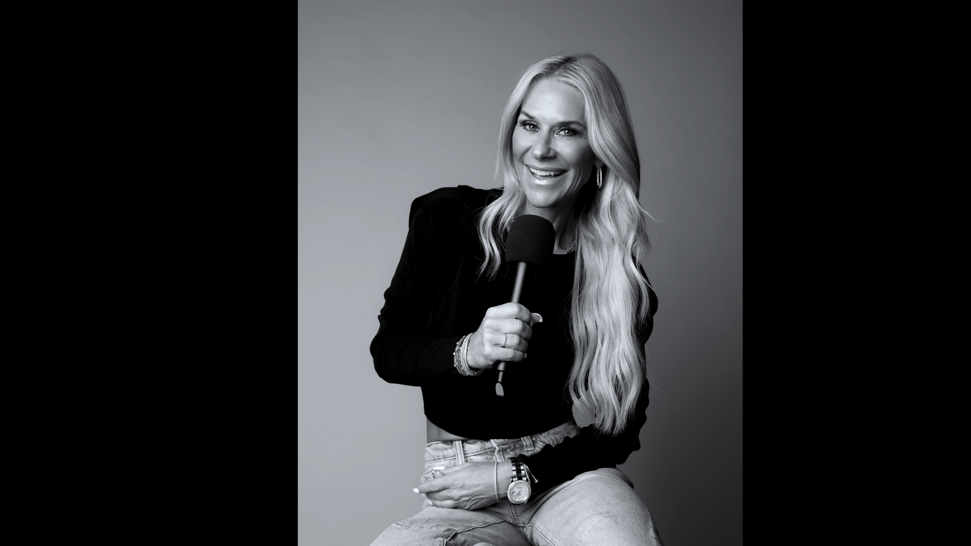 Black and white photo of a woman with long wavy hair holding a microphone, smiling, wearing a black top, jeans, earrings, bracelet, and watch, sitting against a neutral background.