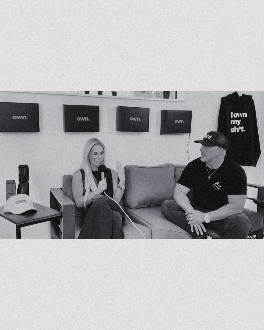Two people sitting on a sofa, one woman speaking into a microphone and a man wearing a cap and black t-shirt with a necklace, in a room with multiple TV screens and merchandise with 'own.' branding.