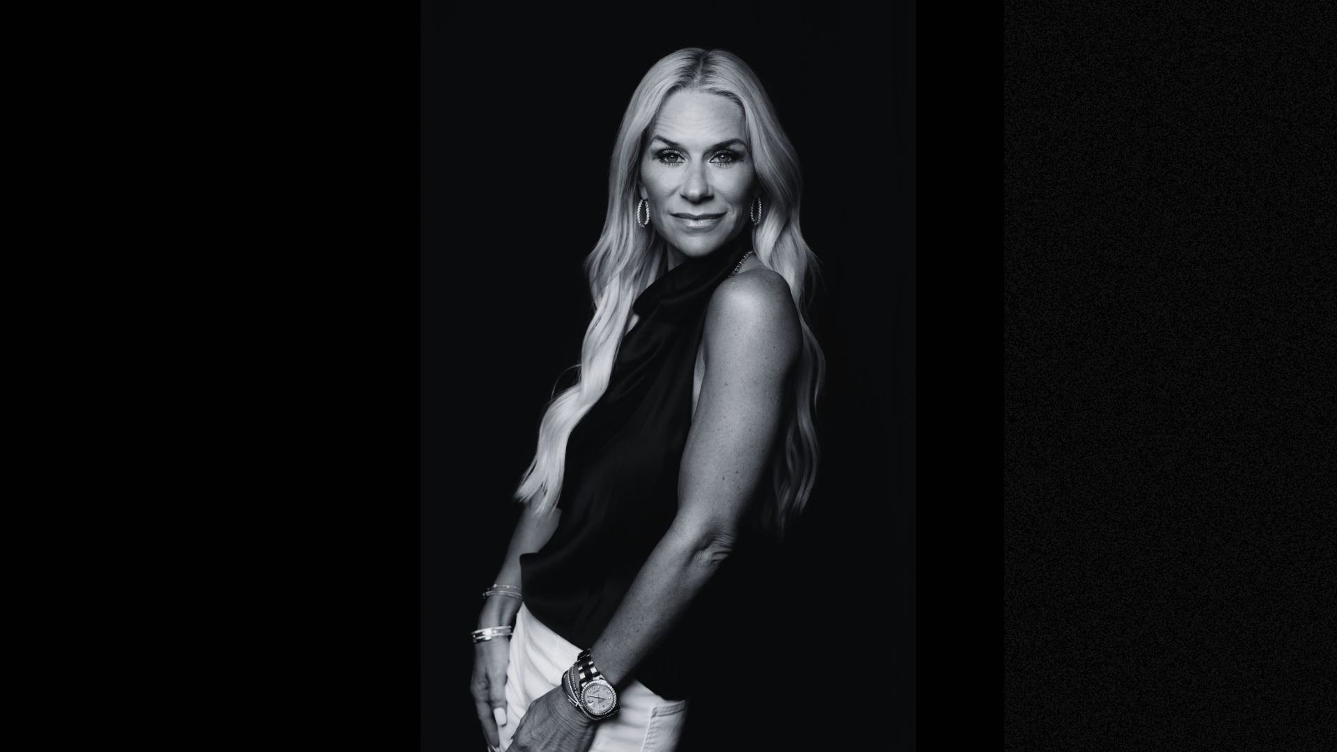 A black and white portrait of a confident woman with long wavy blonde hair, wearing hoop earrings, bracelets, a wristwatch, a sleeveless black top, and white pants, posing against a dark background.