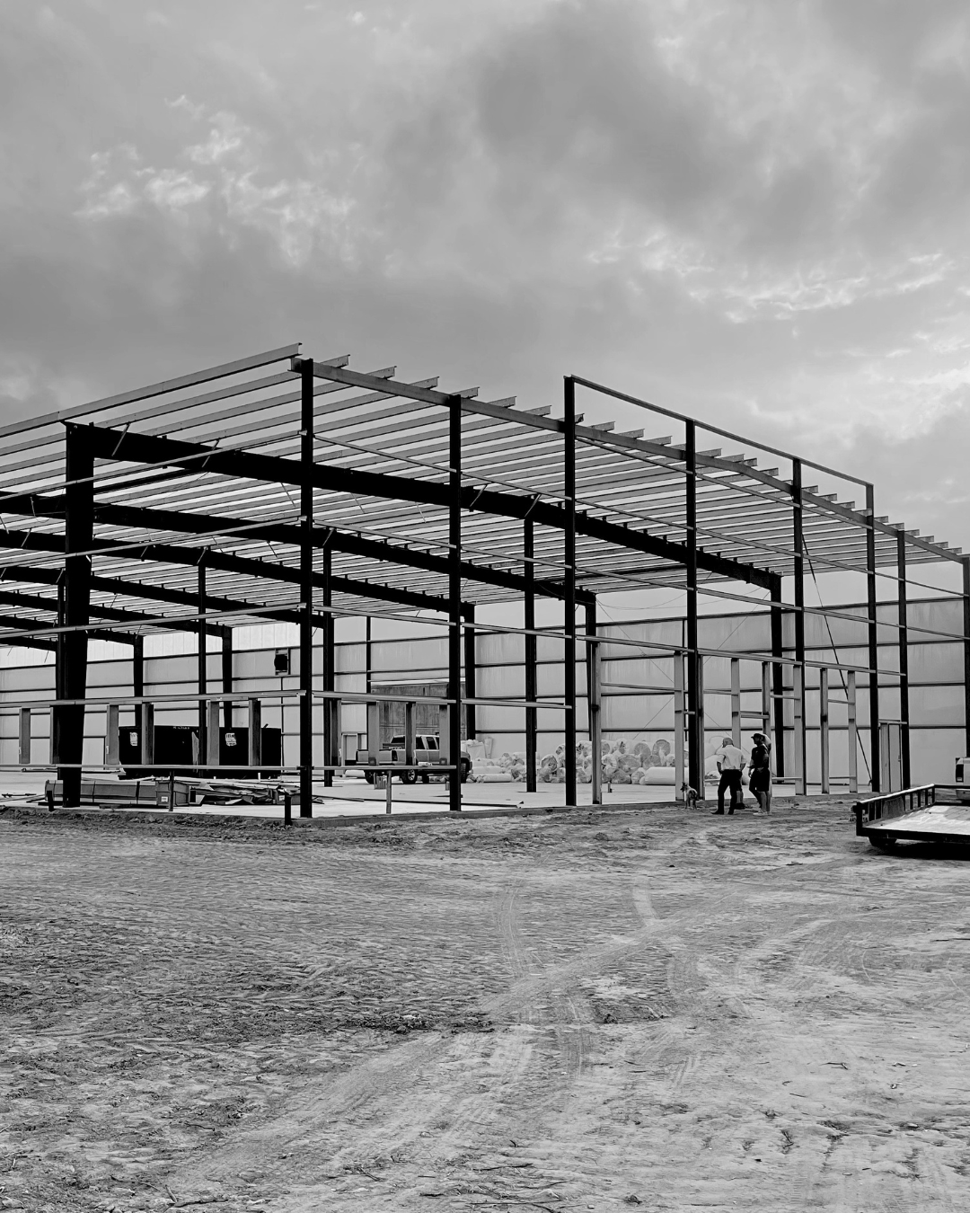 Black and white photo of a building under construction with a visible metal framework, some people standing and talking on the ground nearby, and a cloudy sky overhead.