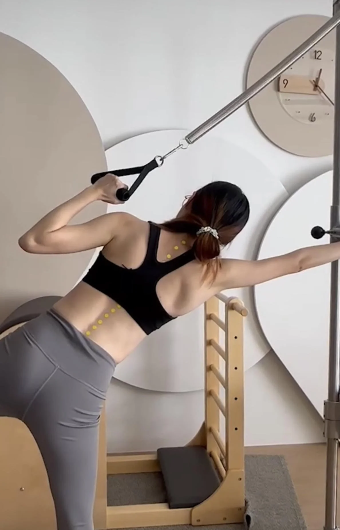 A woman exercising on a resistance machine, stretching her arm in a gym with large wall clocks and circular wall decorations.