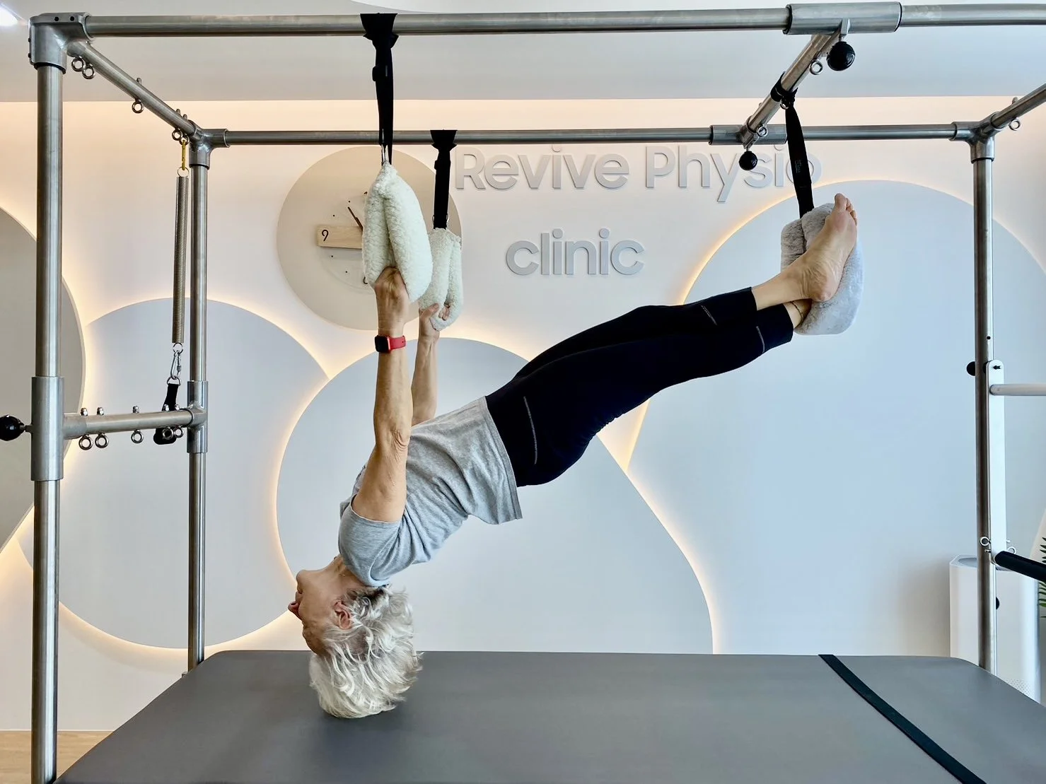 A woman practicing aerial yoga or therapy on a fabric hammock, upside down with her feet secured in foot padding, in a fitness studio labeled 'Revive Physical clinic.'
